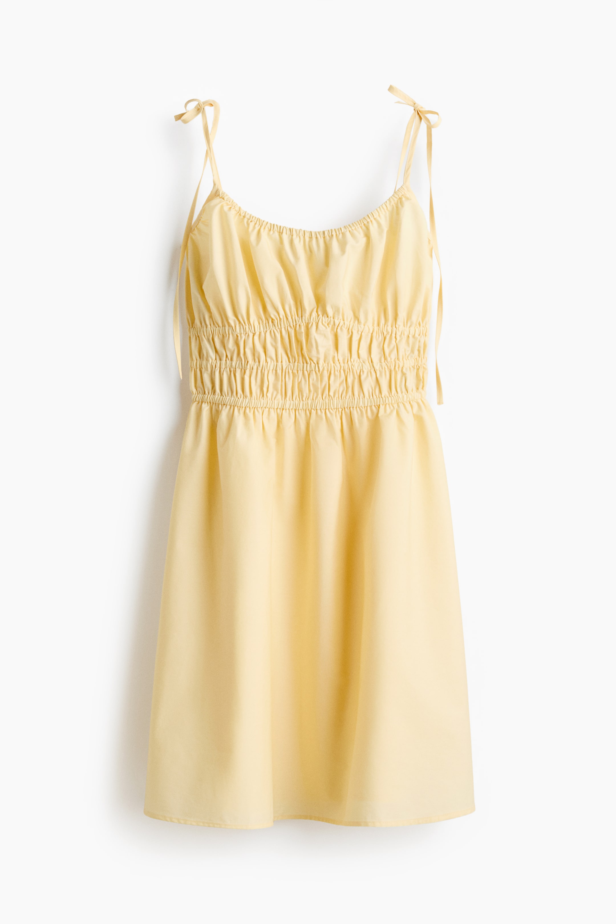 View larger image: A light yellow dress is shown front-facing, with thin tie straps and a gathered neckline. A wide shirred panel shapes the waist, from which a flared skirt extends.