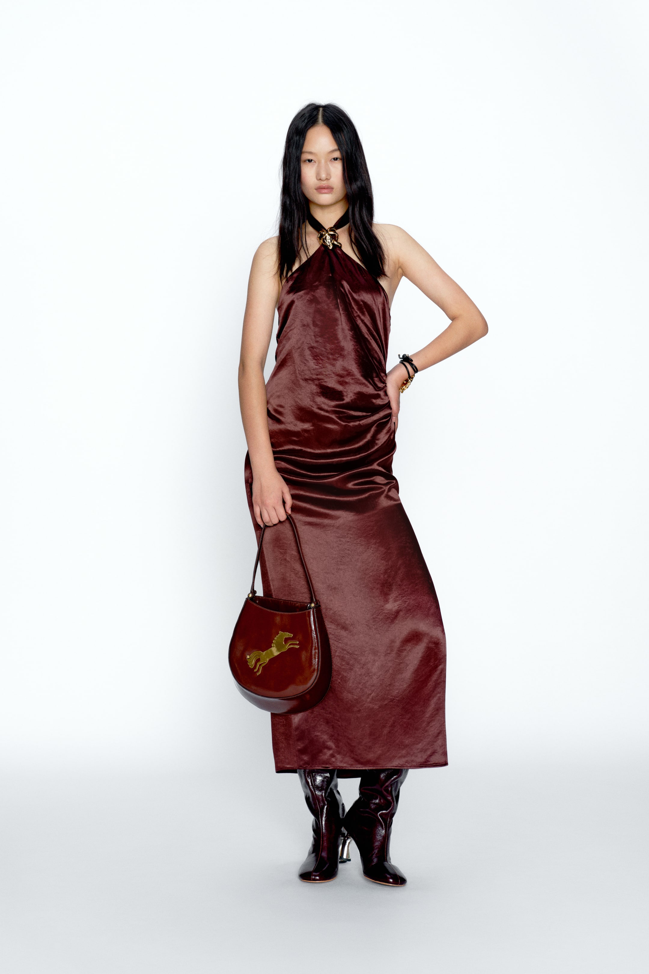 View larger image: A dark reddish-brown midi dress featuring a halter neckline, finished with a gold-toned sculptural clasp securing the straps. The lustrous, lightly textured fabric drapes into a flowing, slip-style silhouette.