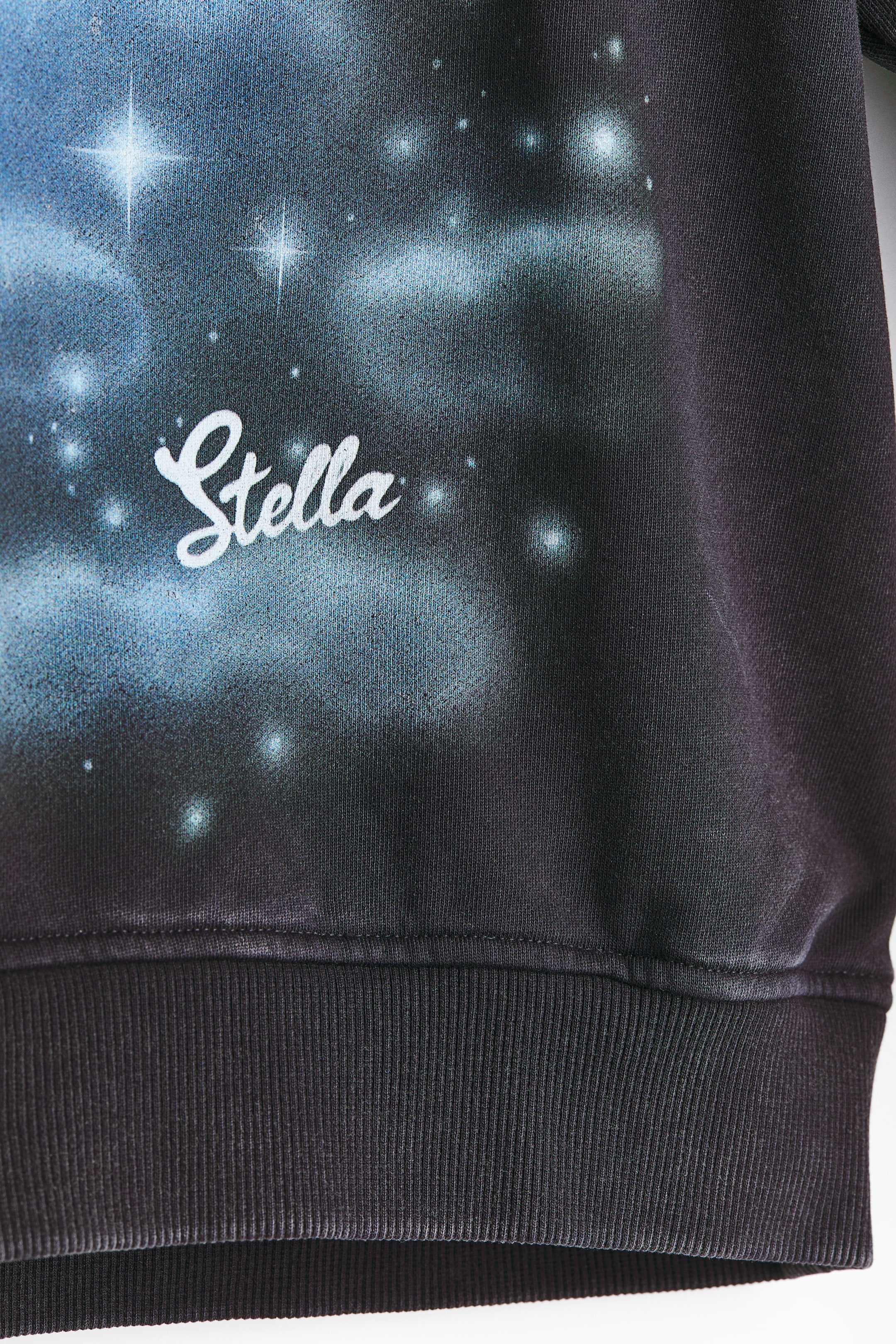 Größeres Bild ansehen: Ein dunkler Hoodie mit einem himmlisch inspirierten Druck auf der Vorderseite, der leuchtend blaue und weiße sternenähnliche Muster sowie den weißen Schriftzug "Stella" zeigt. Der untere Teil der Vorderseite des Hoodies ist mit einem gerippten Saum versehen.