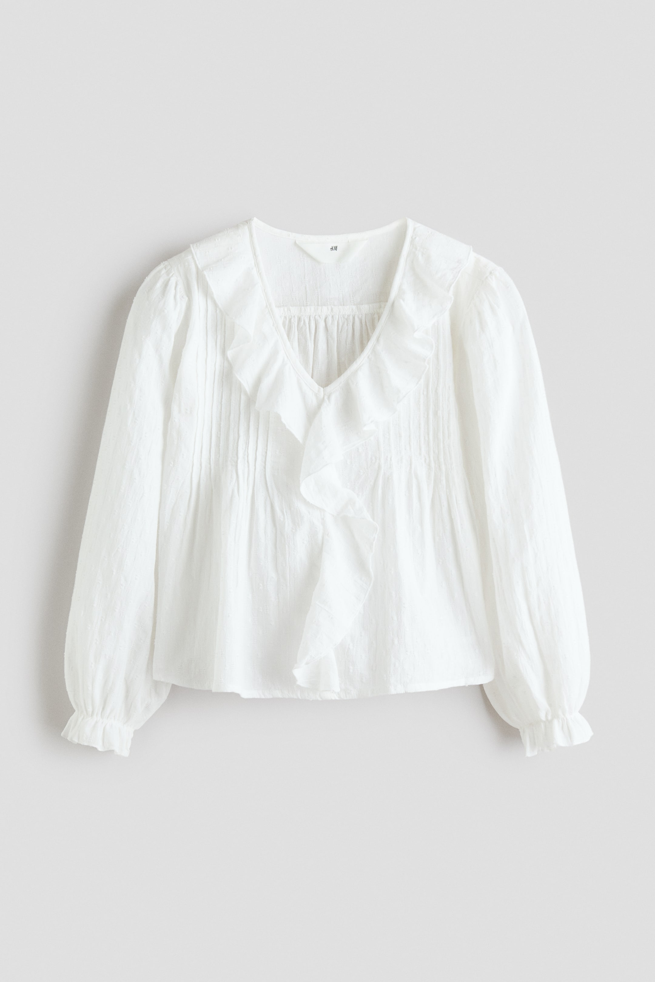 View larger image: The white blouse features a V-neckline accentuated by ruffles, with vertical pintucks gracing the front bodice and a central ruffle detail. The design is completed with long, puffed sleeves that have elasticated, ruffled cuffs.
