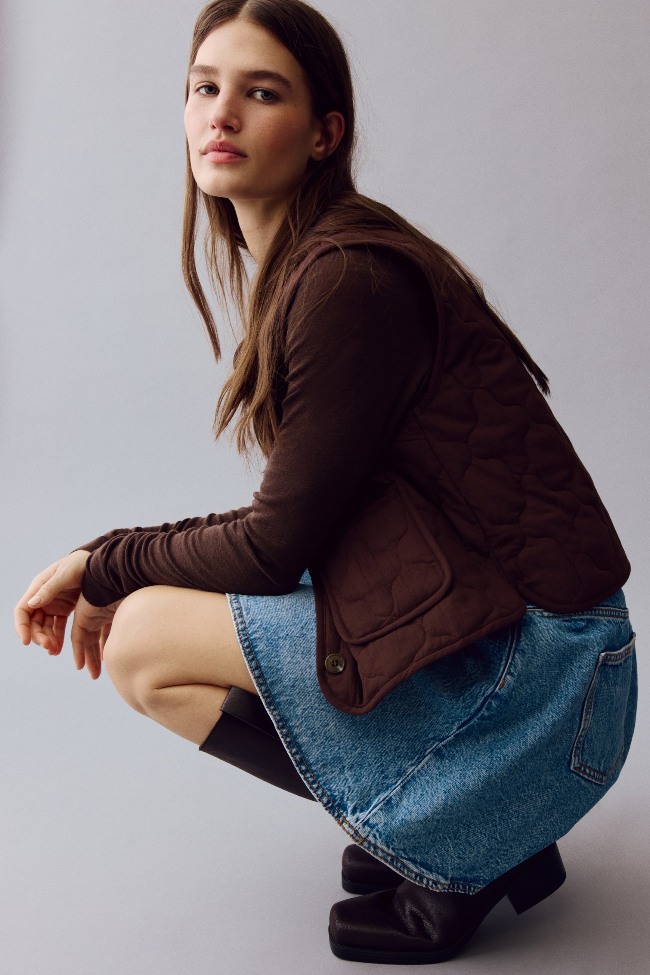 View larger image: A dark brown, fitted, quilted gilet is worn by the model. It features a crinkled texture, binding along the edges, and a patch front pocket with a button closure. The gilet is layered over a long-sleeved top.
