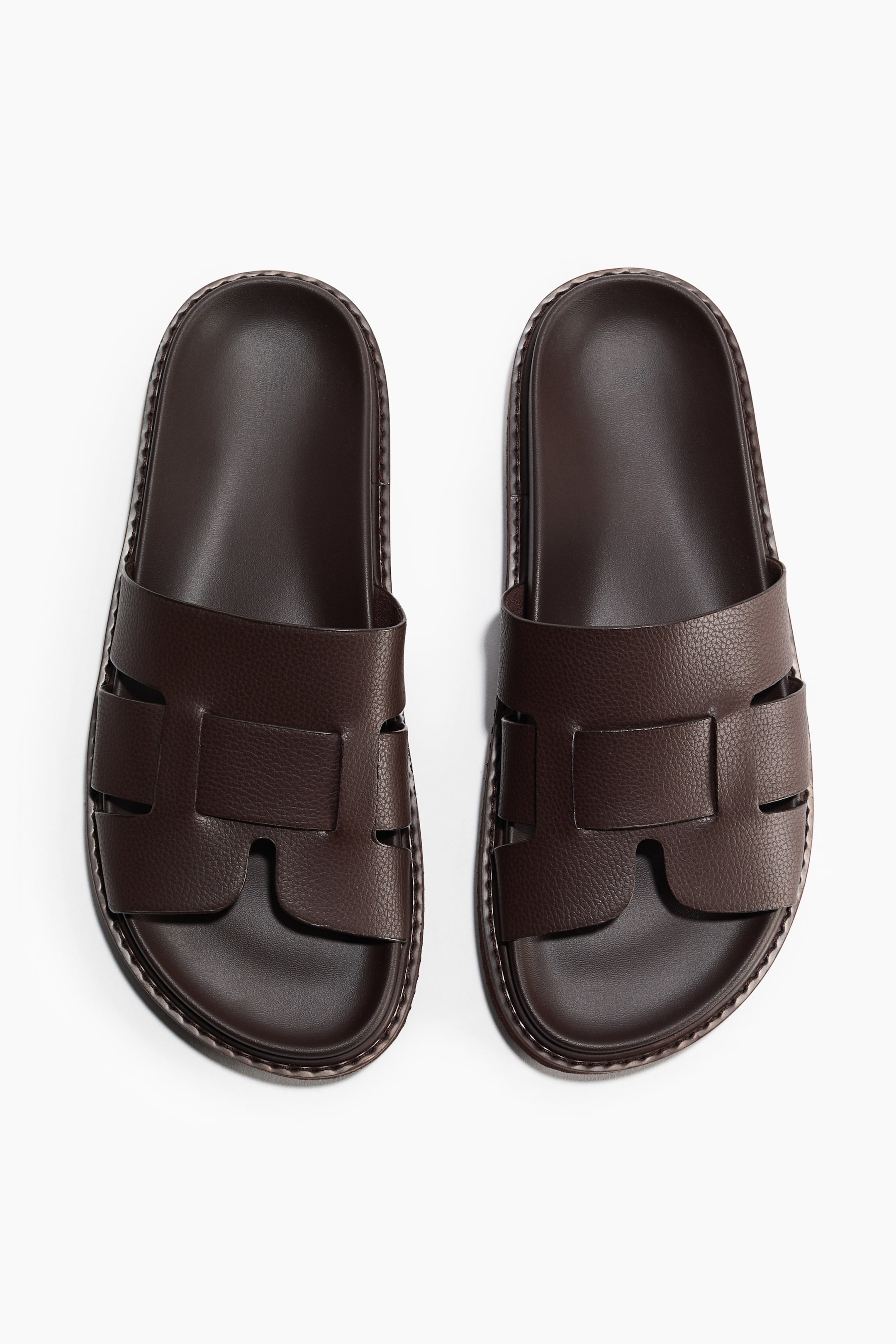 View larger image: A pair of dark brown slide sandals is placed side-by-side, facing forward. Each sandal showcases a wide upper strap made from pebbled material, forming H-shaped cutouts, and a smooth footbed. The thick sole is adorned with visible stitching around its edge.