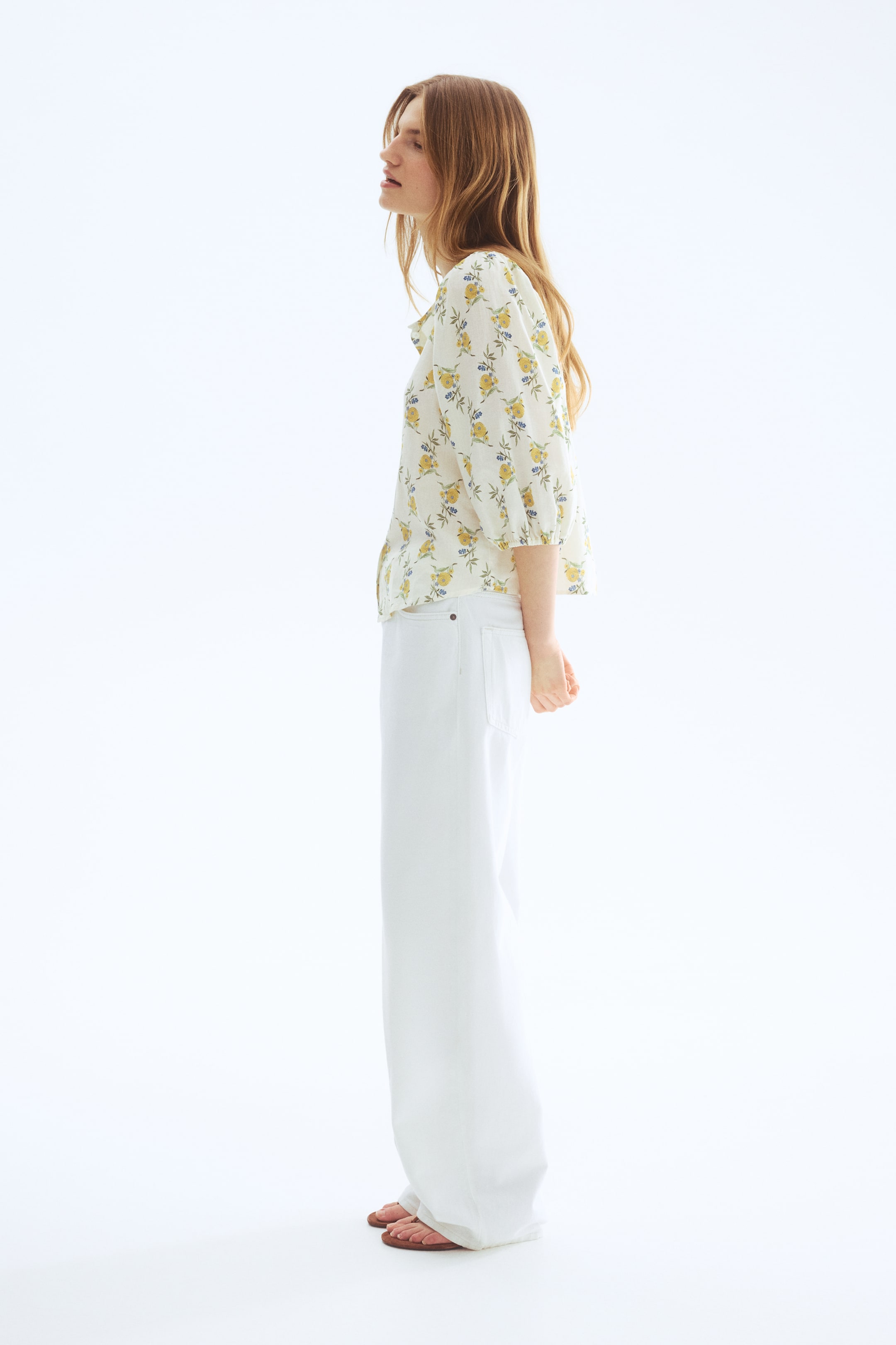 Ver imagem maior: Um conjunto composto por uma blusa branca de mangas curtas com um padrão floral amarelo e detalhes azuis, usado com calças brancas de perna larga e sandálias rasas castanhas, apresentado de um ângulo lateral. A blusa exibe mangas bufantes que terminam acima do cotovelo, enquanto as calças apresentam uma silhueta descontraída e de comprimento total.
