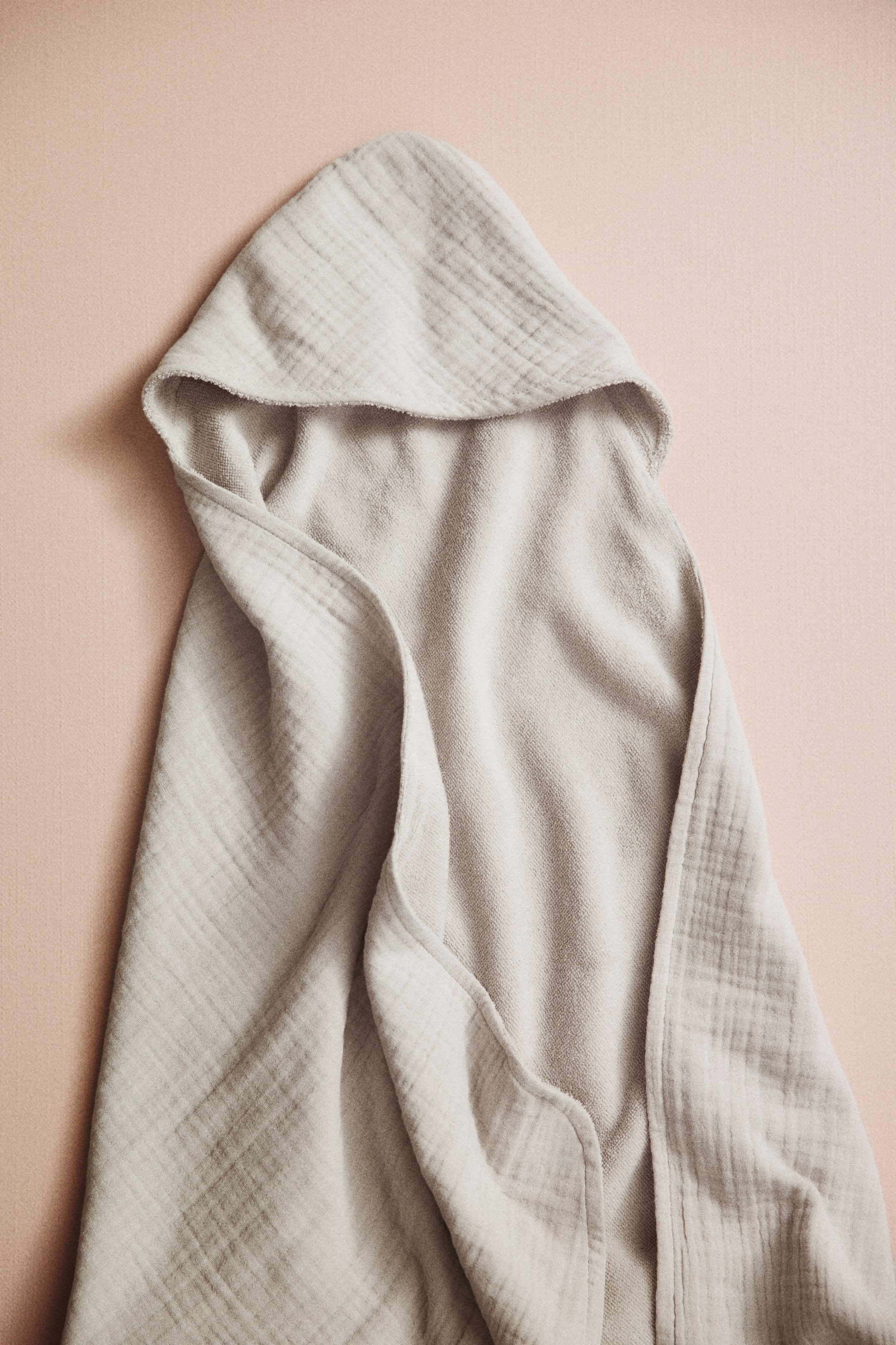 View larger image: A hooded baby towel is laid out flat, highlighting its double-layered design. The outer layer displays a light beige cotton muslin with a crinkled texture, while the inner lining features a soft, uniform terry fabric.