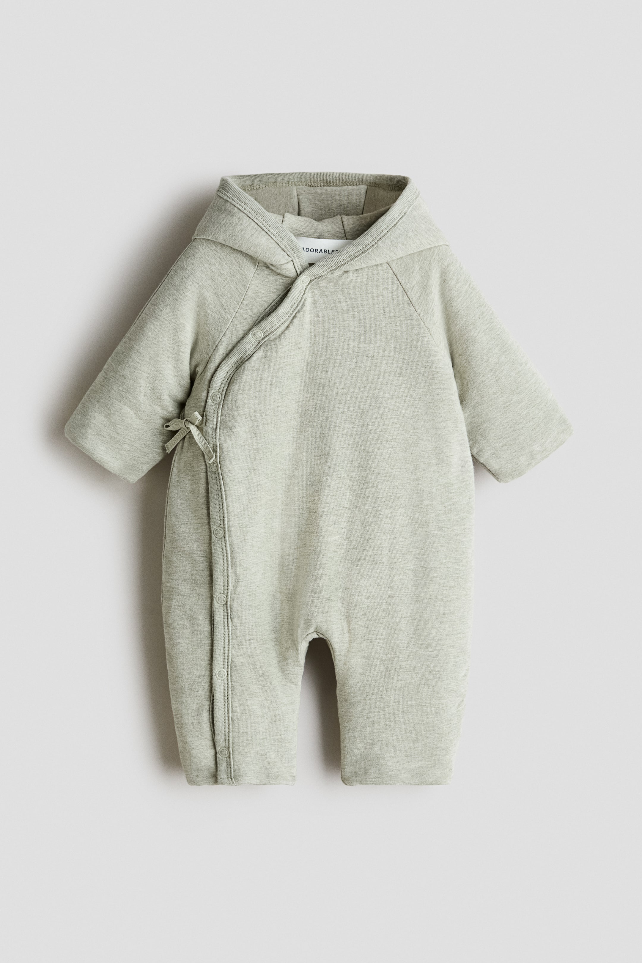 View larger image: A light green padded pramsuit is shown from the front, featuring an attached hood and a soft jersey texture. Its wrapover front fastens with ties and press-studs on the left side, with the press-studs extending down the left leg.