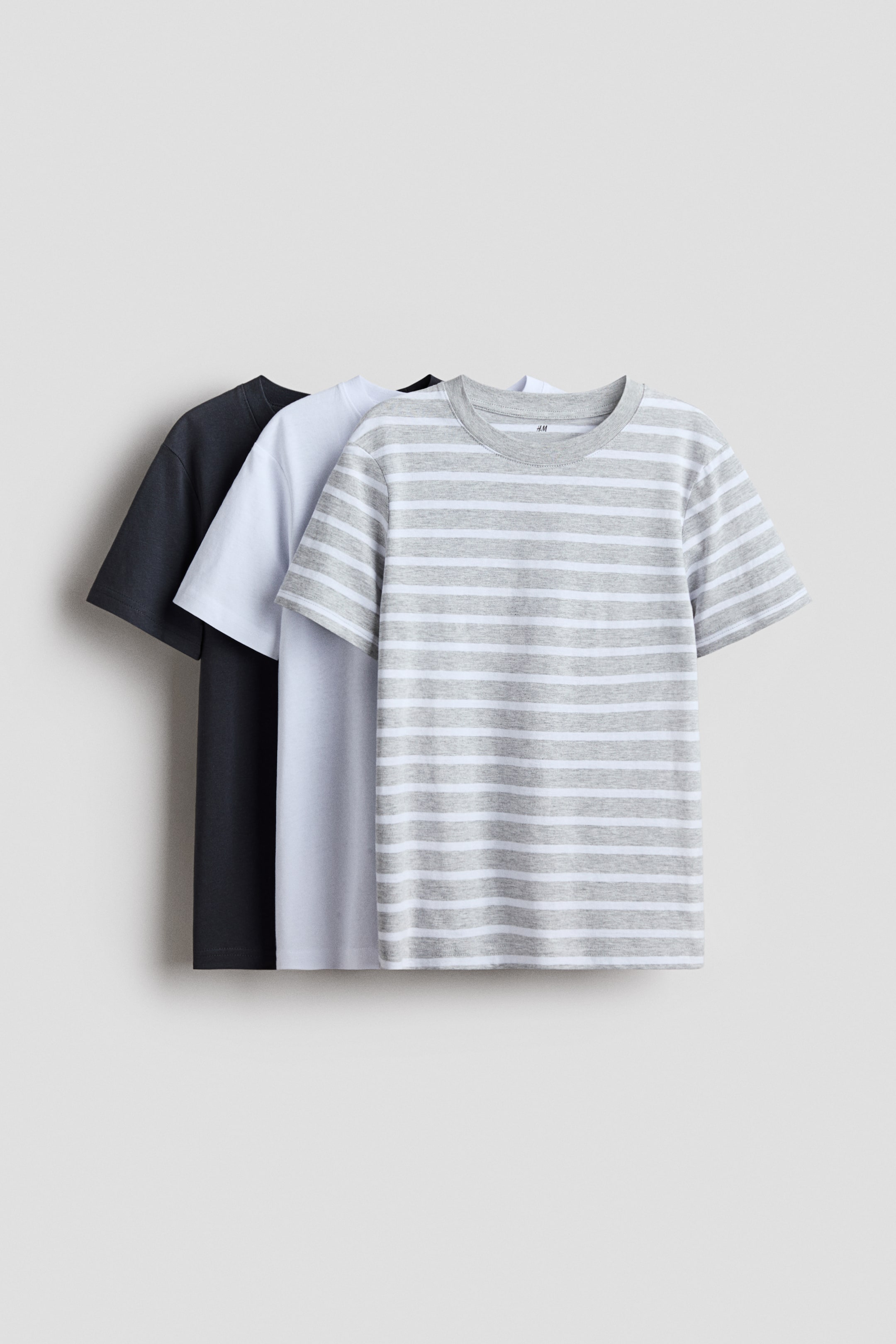 View larger image: Three round-necked T-shirts in soft cotton jersey are displayed with their fronts visible, layered one behind the other. The front T-shirt features grey and white horizontal stripes and a small brand label on its inner back neckband, while a solid white T-shirt and a solid dark grey T-shirt are layered behind it.