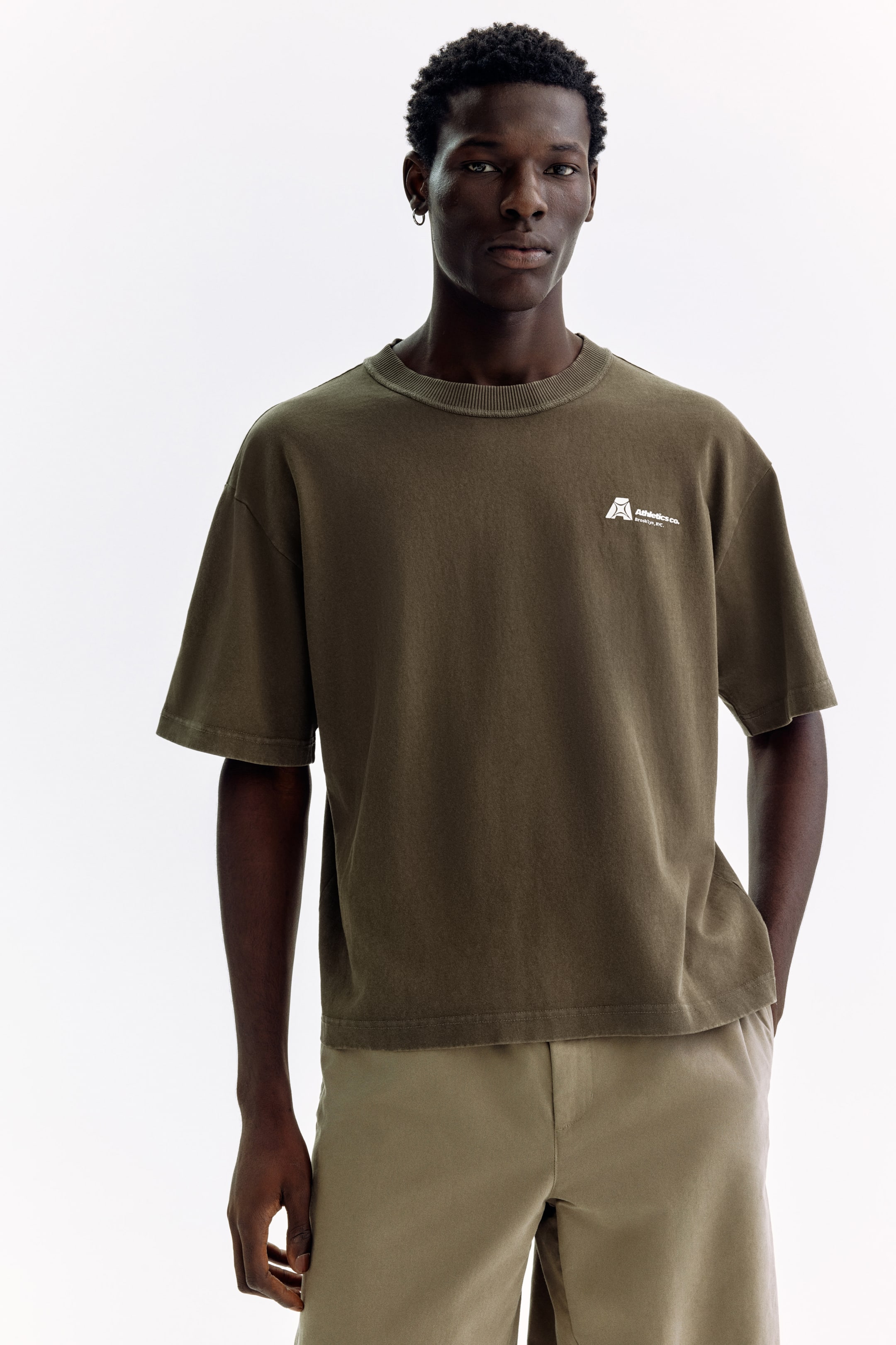 View larger image: An olive green t-shirt is showcased, featuring a ribbed crew neck, short sleeves, and a straight hem. A small white logo, displaying a stylized 'A' above 'Athletics Co. Brooklyn, NY,' appears on the left chest, with the t-shirt offering a relaxed, oversized fit.