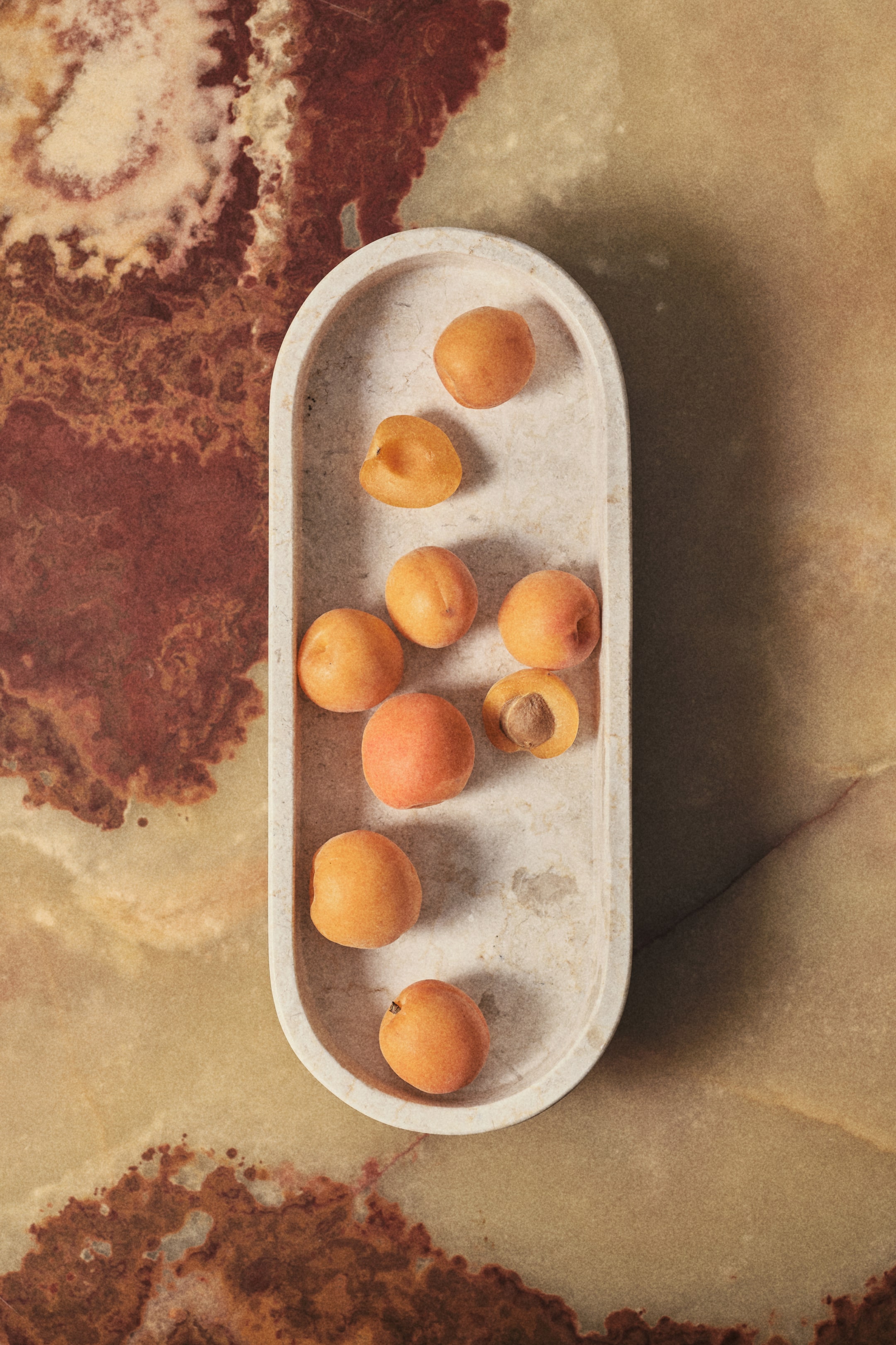 Voir une image plus grande: Une assiette ovale, fabriquée en marbre de couleur claire, est surélevée sur un pied, présentant une silhouette raffinée. Sa surface en marbre montre des variations naturelles de couleur et un veinage subtil, et elle contient plusieurs abricots.
