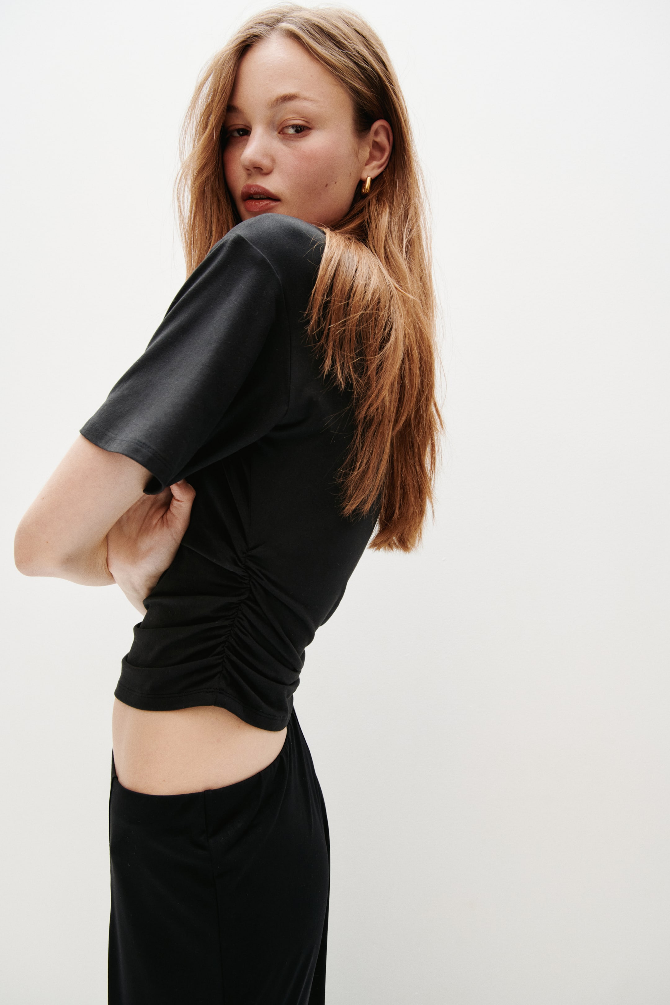 הצגת תמונה גדולה יותר: הדוגמנית לובשת חולצת טריקו שחורה. החולצה כוללת כתפיים נופלות, כיווצים בצדדים שיוצרים סילואט מחוטב עם אפקט רך ומתנפנף, ושוליים קצרים שחושפים את הבטן.