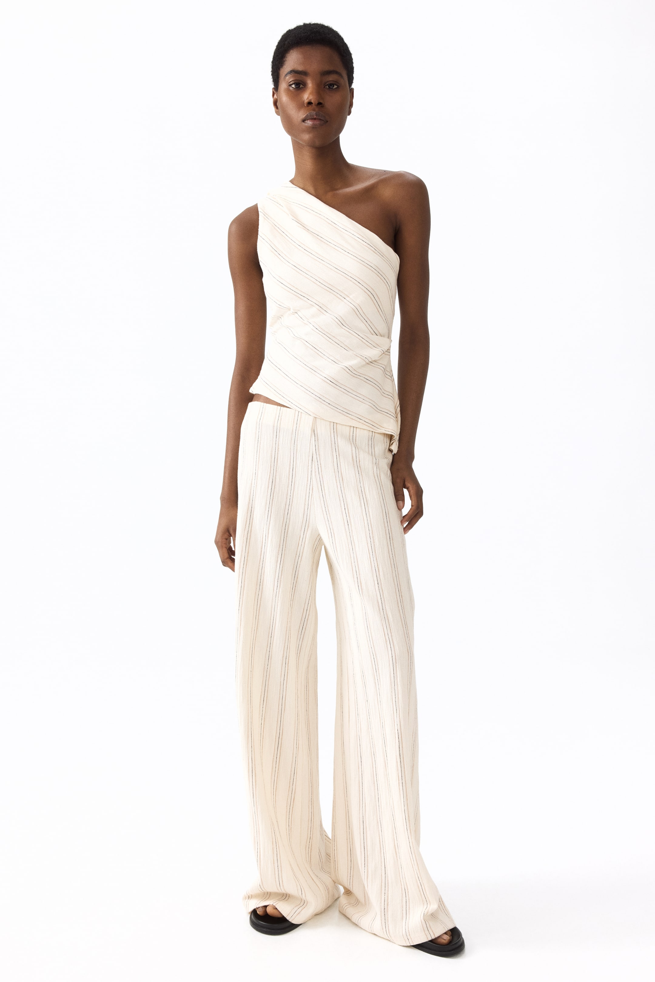 Ingrandisci l'immagine: I pantaloni in cotone strutturato indossati dalla modella presentano un colore crema, una finitura strutturata, sottili righe verticali e una silhouette a gamba larga. La modella li abbina a un top monospalla coordinato.