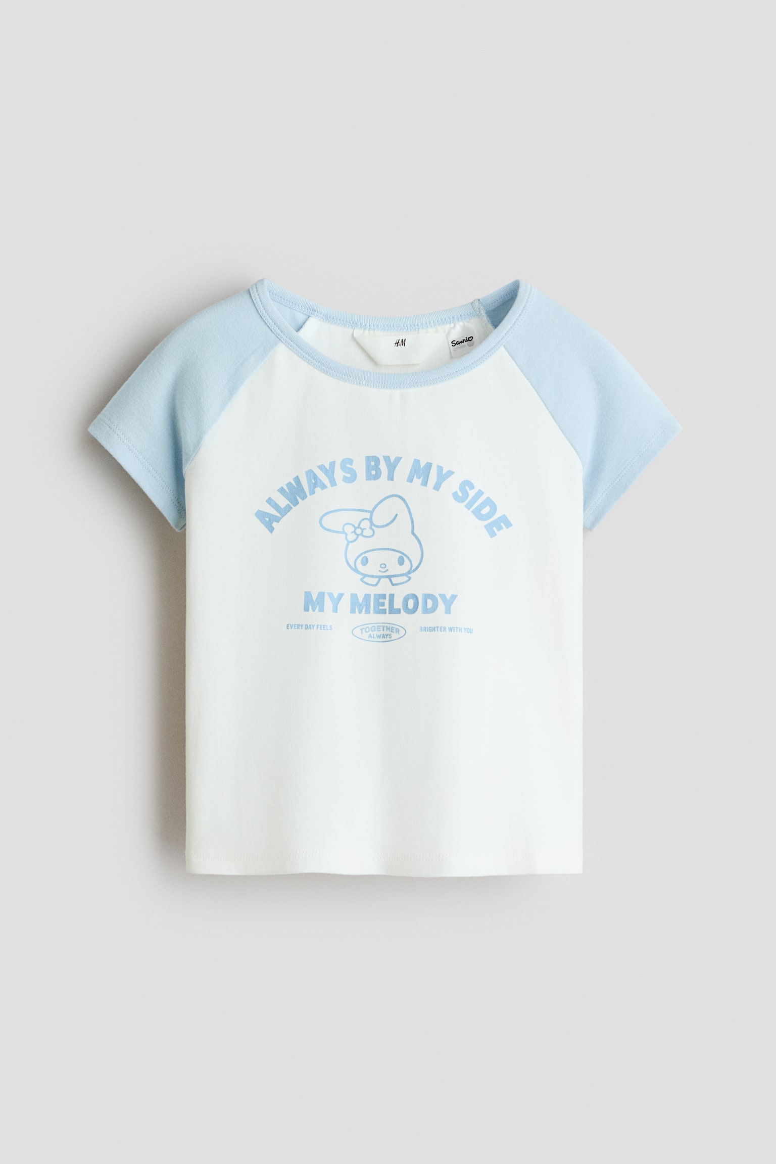 Un T-shirt en jersey de coton blanc est prÃ©sentÃ© de face, avec de courtes manches raglan bleu pÃ¢le et un col assorti Ã  bord-cÃ´te. Le devant au centre affiche un graphique bleu pÃ¢le du personnage My Melody avec le texte imprimÃ© Â« ALWAYS BY MY SIDE Â» et Â« MY MELODY Â».