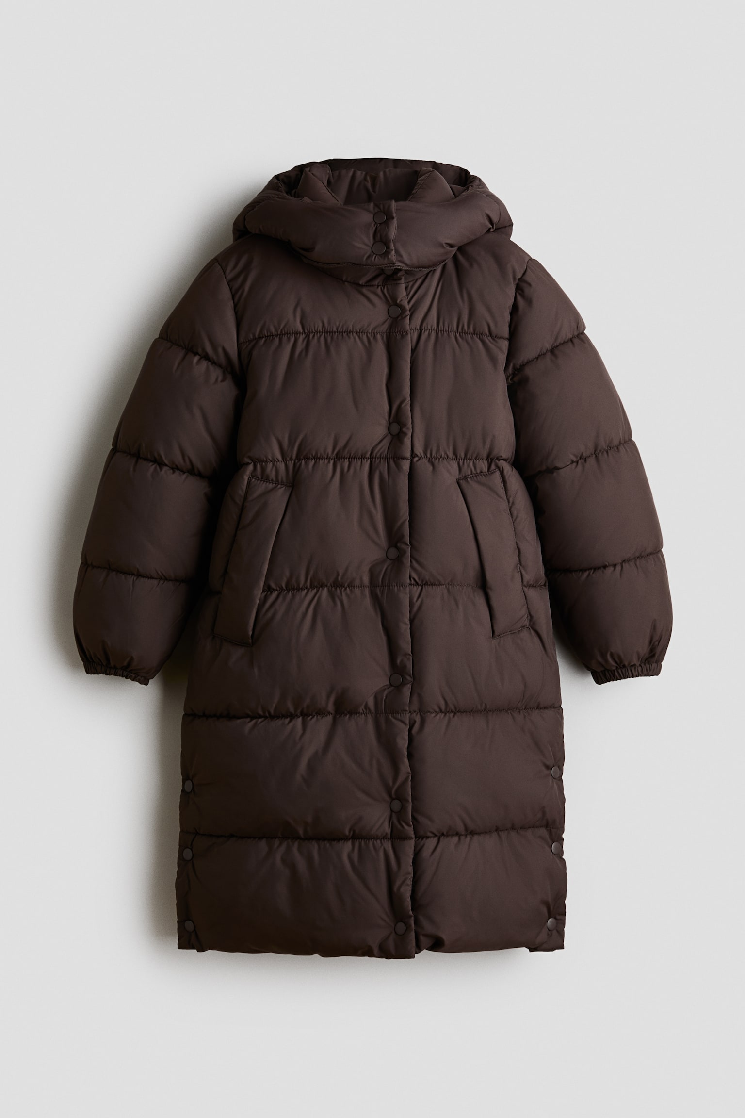 Eine lange, dunkelbraune, gepolsterte Pufferjacke aus gestepptem Stoff ist von vorne zu sehen. Sie verfÃ¼gt Ã¼ber einen hohen Kragen mit angesetzter Kapuze, eine Windschutzleiste mit DruckknÃ¶pfen und schrÃ¤ge Paspeltaschen. Die Jacke hat lange Ãrmel mit elastischen BÃ¼ndchen sowie seitliche Schlitze mit DruckknopfverschlÃ¼ssen nahe dem Saum.