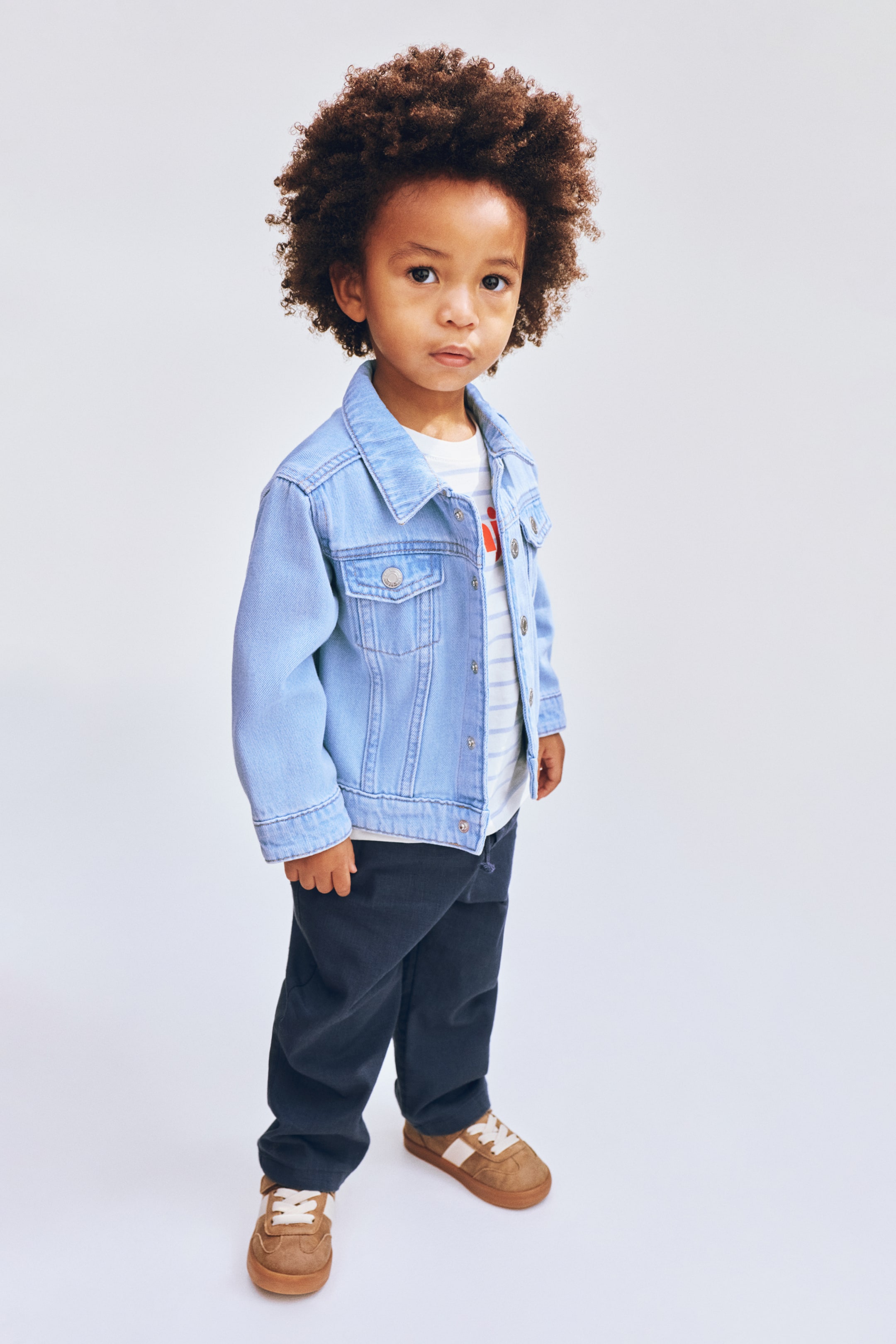 View larger image: A light blue denim jacket featuring a pointed collar and two chest flap pockets with snap buttons. The jacket is styled over a white and blue striped t-shirt, with its front snap buttons open.