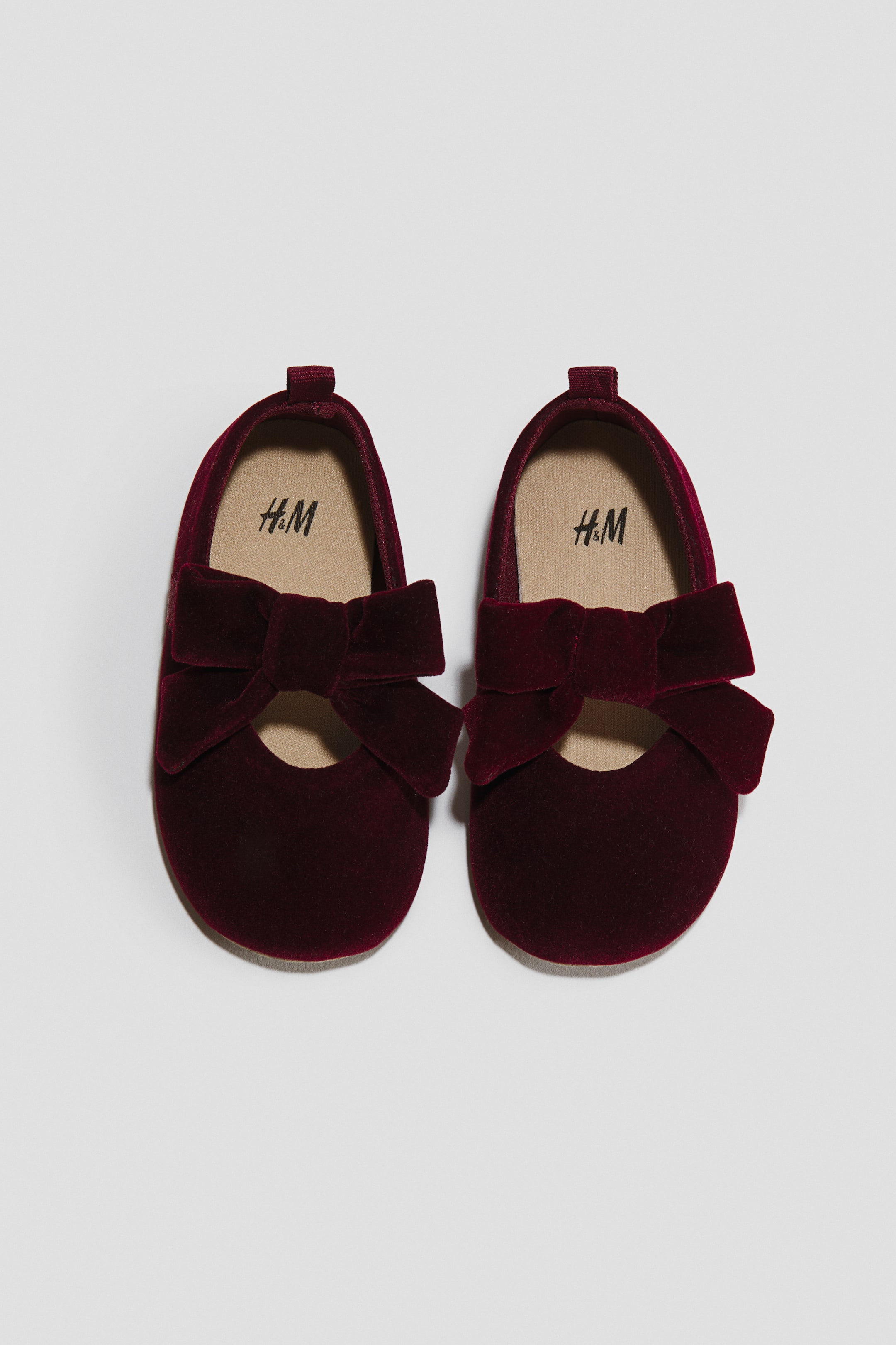 View larger image: A pair of burgundy velvet Mary Jane shoes is facing front, showcasing a large matching velvet bow detail across the instep. The shoes feature a rounded toe design, a small heel pull tab, and a contrasting beige inner lining.