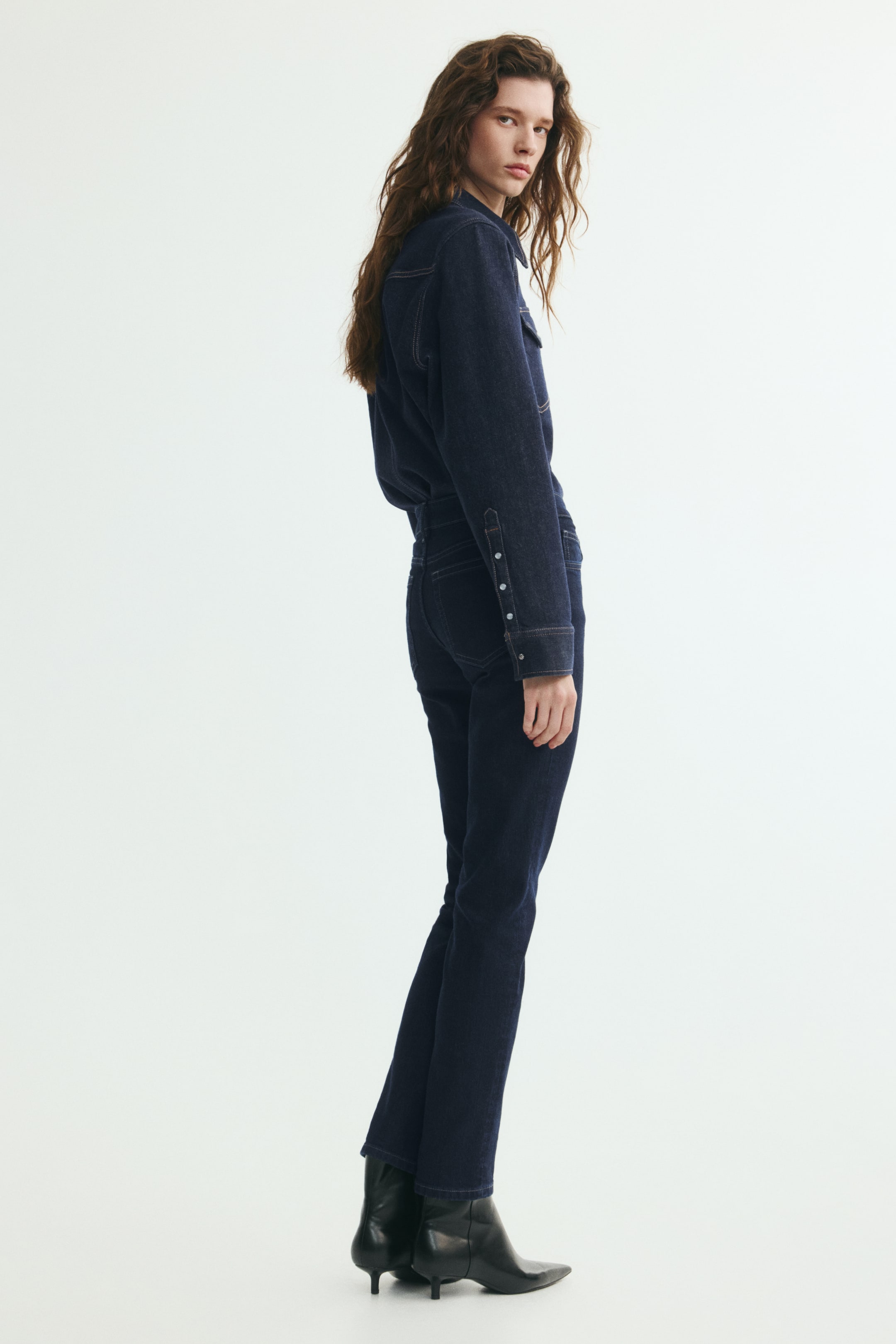 Xem ảnh lớn hơn: Quần jeans denim xanh đậm dáng ôm, cạp vừa được người mẫu mặc, được chụp từ góc nghiêng. Quần được phối cùng áo sơ mi denim xanh đậm đồng bộ được sơ vin và bốt cao gót mũi nhọn màu đen.