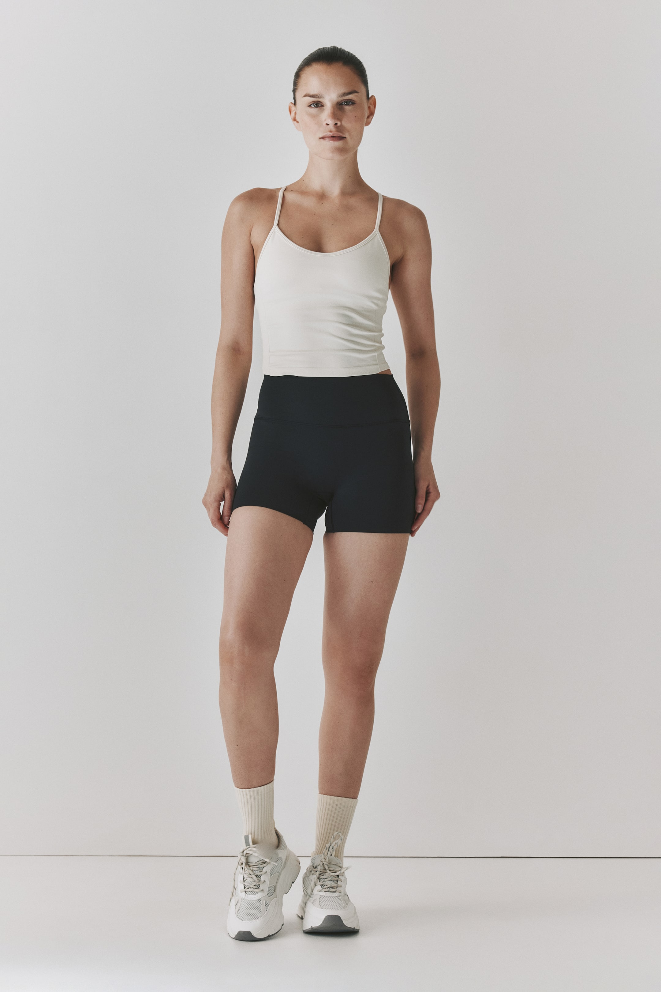 Grotere afbeelding bekijken: Een off-white naadloze sporttop met DryMove?, met dunne spaghettibandjes en een kortere zoom, wordt gedragen door het model, die het combineert met zwarte hooggetailleerde shorts.