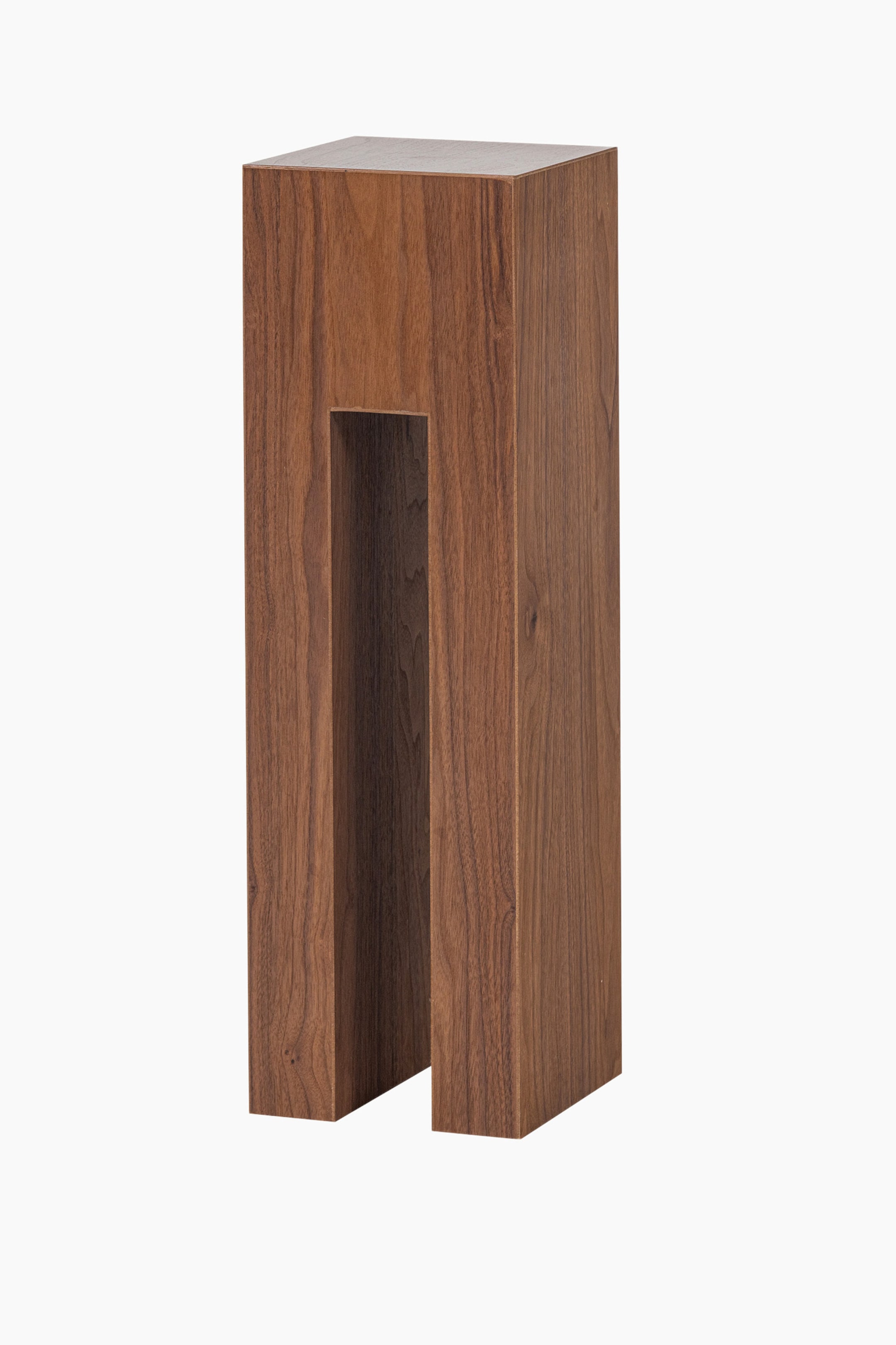 Größeres Bild anzeigen: Ein hoher, rechteckiger Holzsockel wird aus einem leicht frontalen rechten Winkel gezeigt und präsentiert eine dunkelbraune Oberfläche mit sichtbarer Holzmaserung. Ein markanter, vertikaler rechteckiger Ausschnitt erstreckt sich von der Basis nach oben auf der Vorderseite.