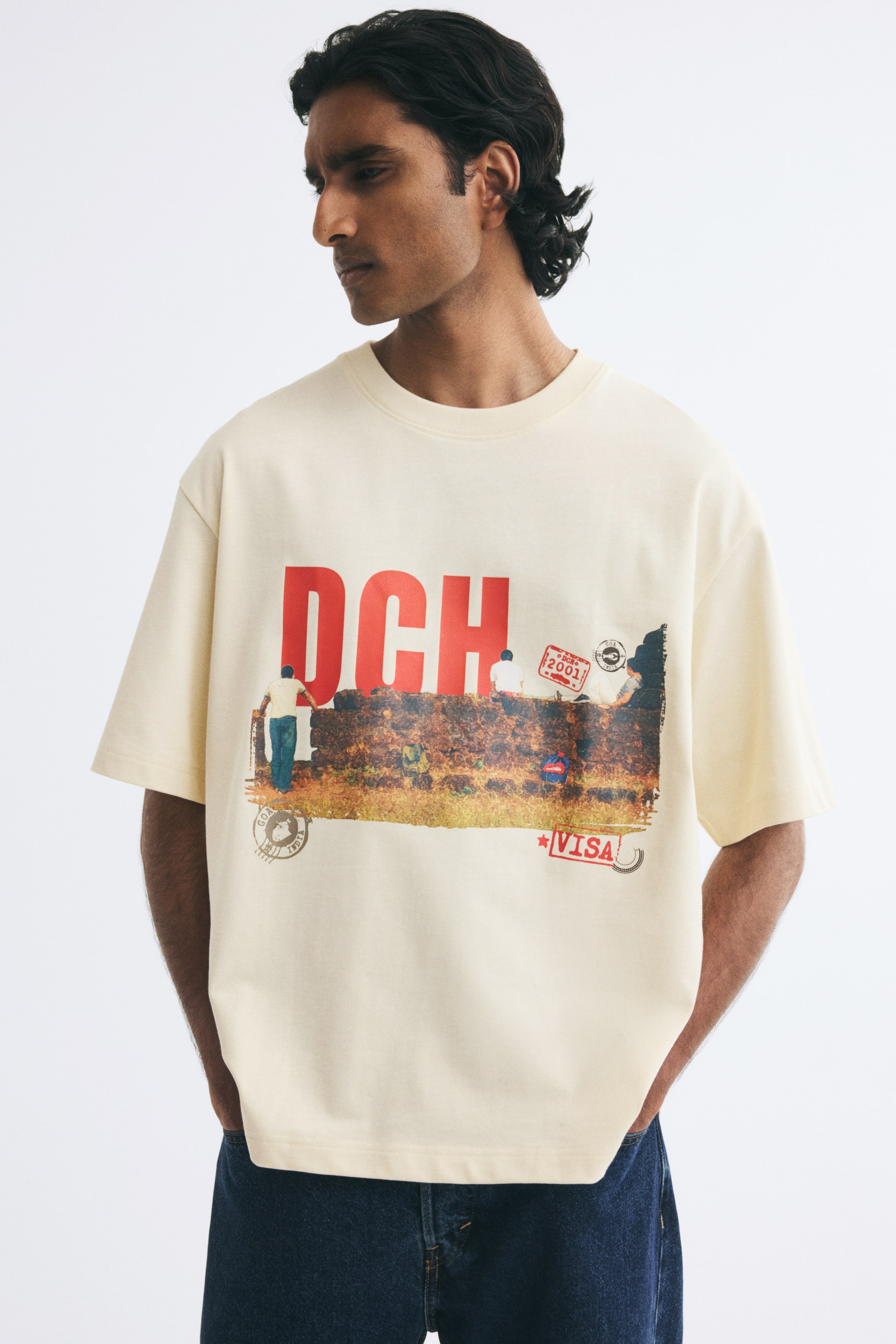 View larger image: The model is wearing a cream-coloured T-shirt in heavyweight cotton jersey, paired with dark blue jeans. The relaxed-fit tee features dropped shoulders, a ribbed crew neck, and a large front graphic print with the red letters "DCH" and stamp motifs.