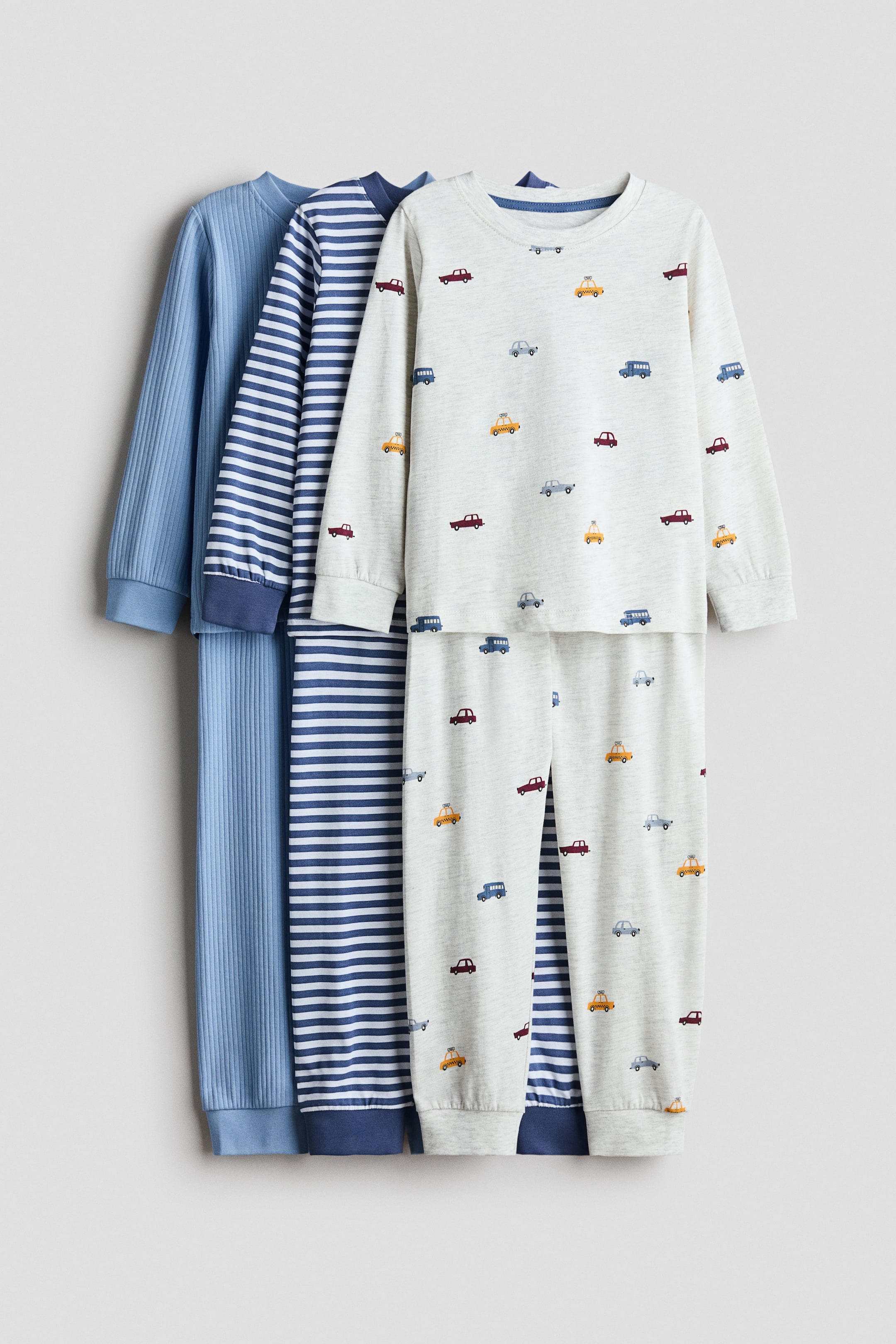 Büyük resmi görüntüle: Üç adet yumuşak pamuklu jarse pijama takımı sunulmaktadır. Her biri yuvarlak ve ribana detaylı yakalı, uzun ve manşetli kollu üst ile lastikli bel ve manşetli paçalı altlardan oluşmaktadır. İlk takım, nervürlü dokusuyla açık mavi renkte, ikinci takım lacivert ve beyaz çizgili, üçüncü takım ise renkli araba desenli açık gri renktedir.