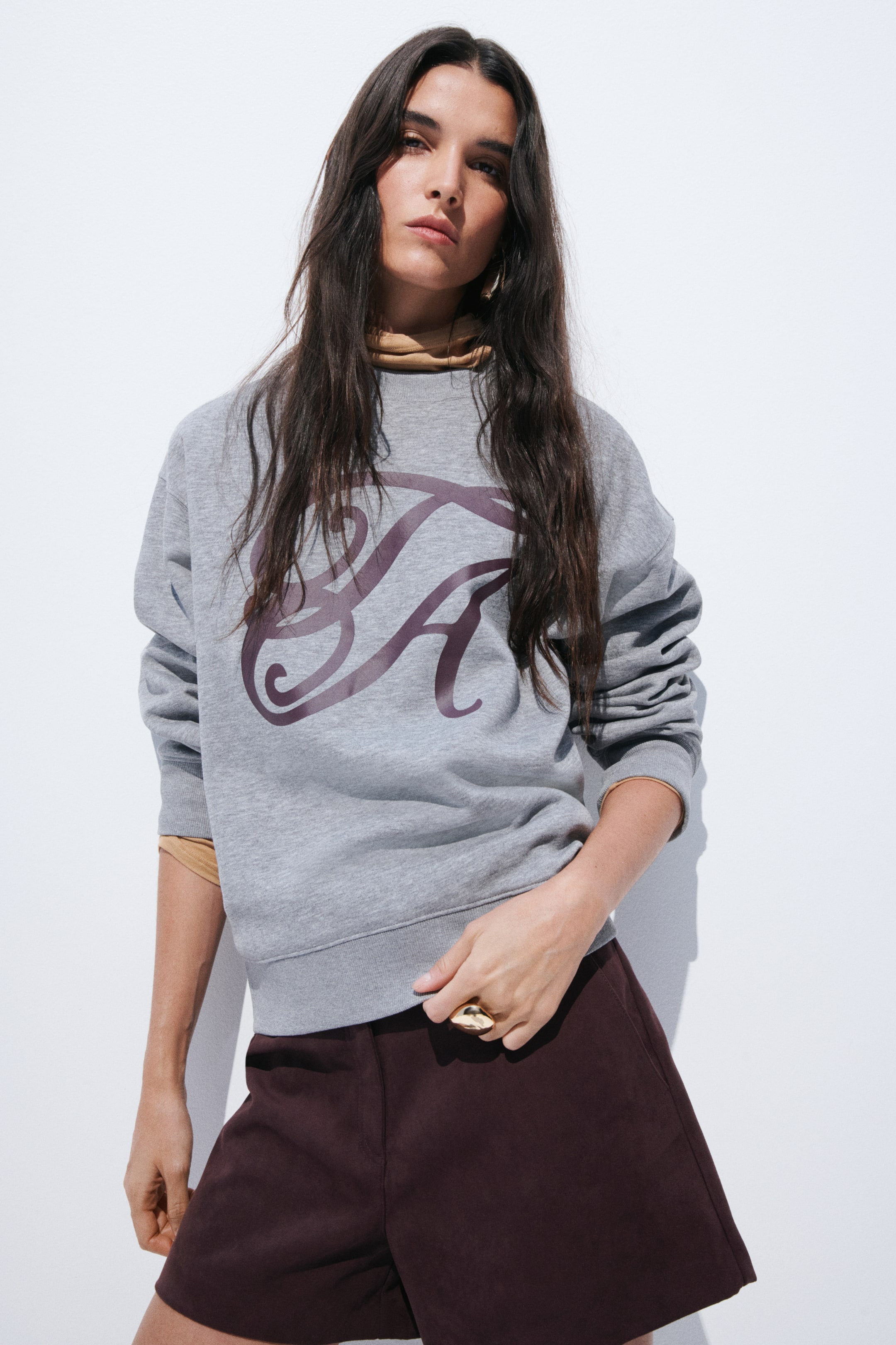 View larger image: A grey sweatshirt featuring a large, dark purple, stylised "SA" graphic on the front. It displays a relaxed fit with a ribbed hem, and a tan long-sleeve top is layered underneath, visible at the neckline and cuffs.