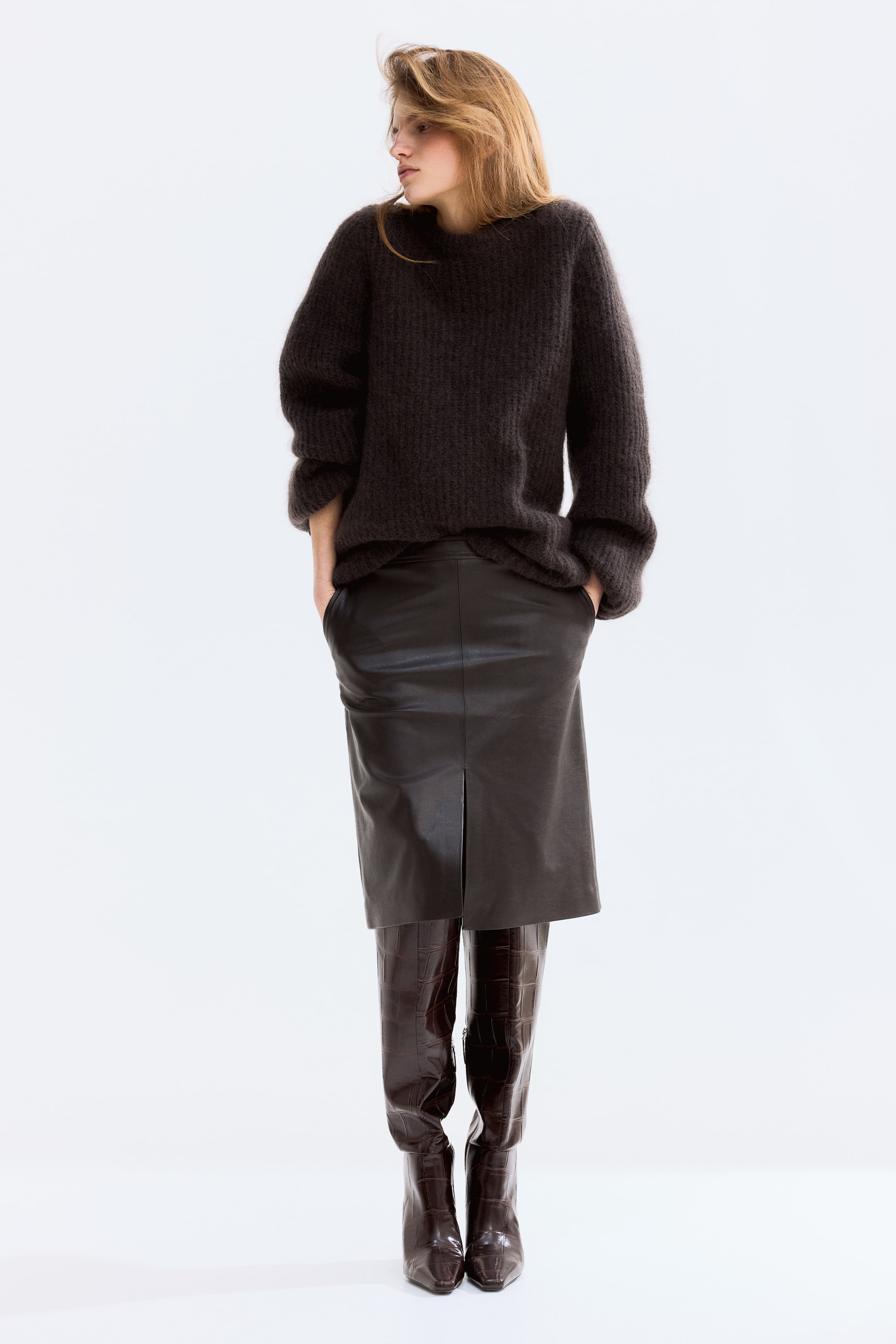 Visa större bild: En mörkbrun luddig stickad tröja med långa, överdimensionerade ärmar bärs över en mörkbrun lädermidi-kjol med en framventil, vilket avslöjar mörkbruna krokodilpräglade knähöga stövlar nedanför, med hela outfiten vänd något åt höger.