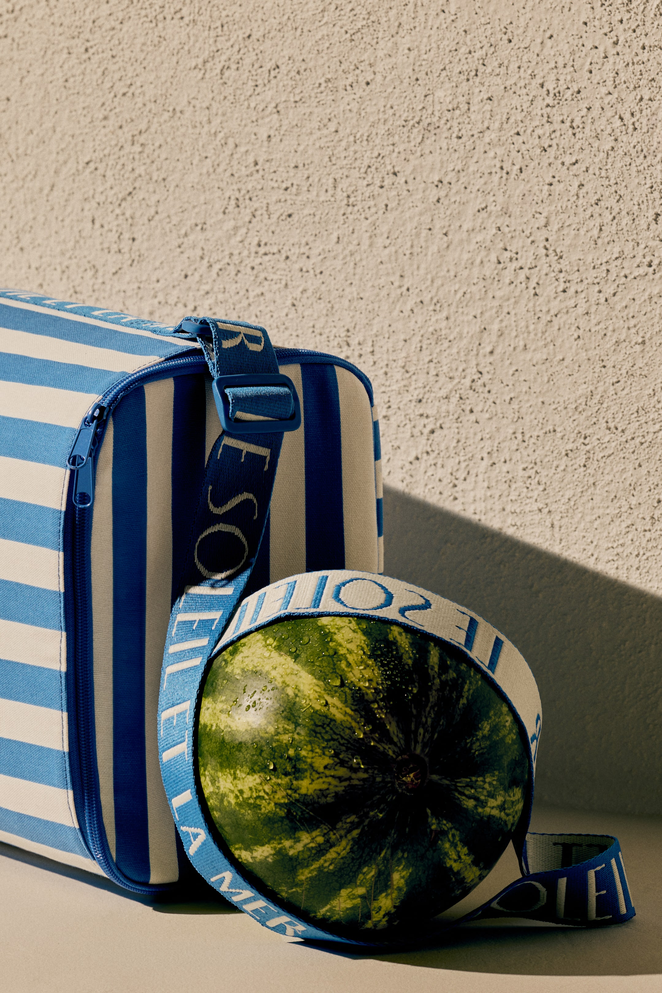 Agrandir l'image: Un sac rectangulaire présente des rayures bleues et blanches, légèrement orienté vers la droite. Une fermeture éclair bleue longe son bord supérieur, et une sangle en tissu bleu avec le texte blanc 'SOIE LEI' entoure une pastèque entière avec des gouttes d'eau.