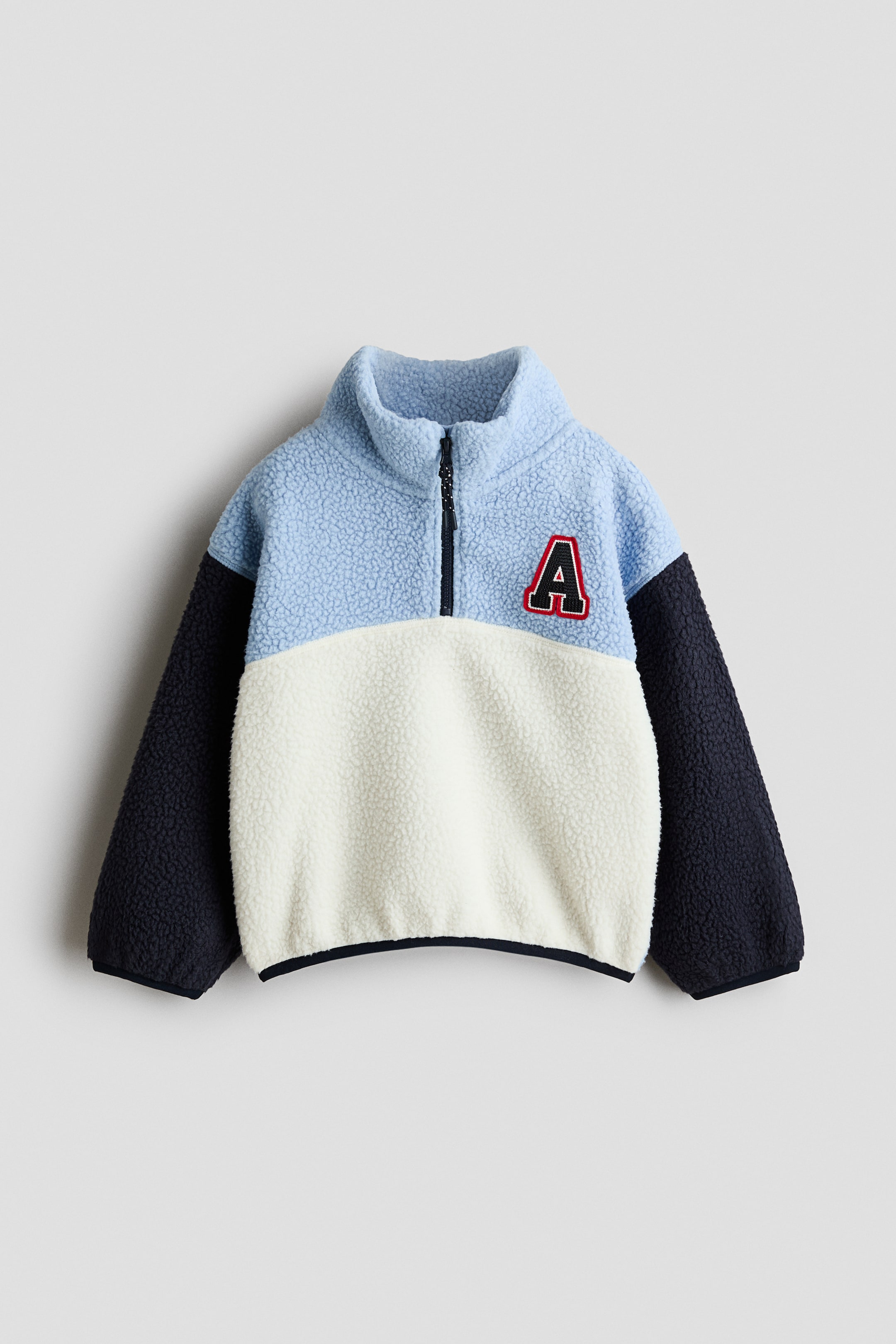 View larger image: A front-facing sherpa fleece top showcasing a colour-blocked design with a light blue stand-up collar and upper body, an off-white lower body, and dark blue long sleeves. A black quarter-zip closure runs down the front, and a red letter "A" patch with white lettering and a black outline is positioned on the left chest, while dark blue binding finishes the cuffs and bottom hem.