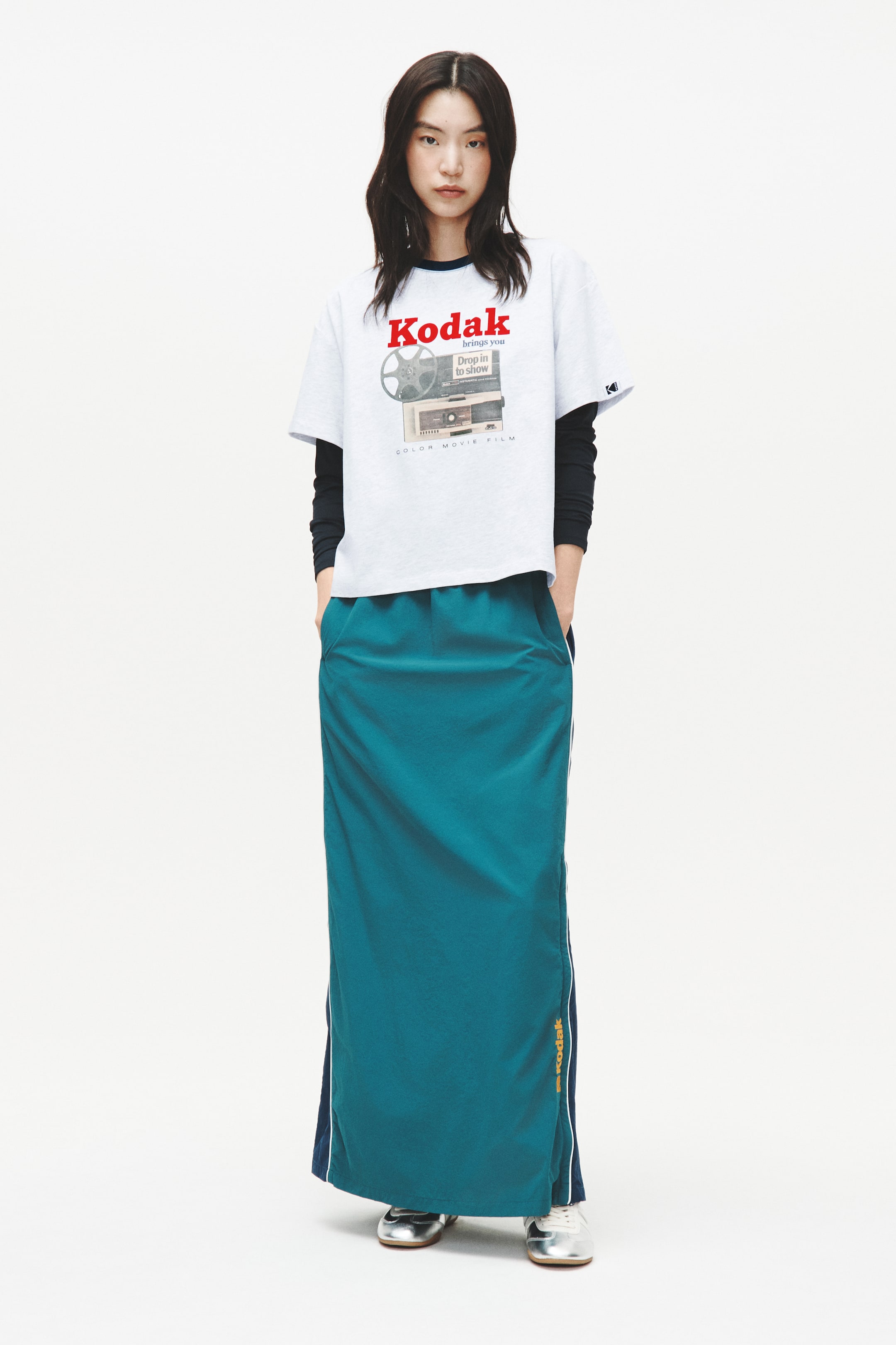 View larger image: A grey heather t-shirt, featuring a red "Kodak" logo and a vintage film projector graphic, is worn over a black long-sleeved shirt, showcasing a dark blue crew neck trim and a small logo on its left sleeve. A teal maxi skirt features an elasticised waistband, side pockets, and white stripes running down the sides, revealing dark blue fabric and a yellow "Kodak" logo on the right side.