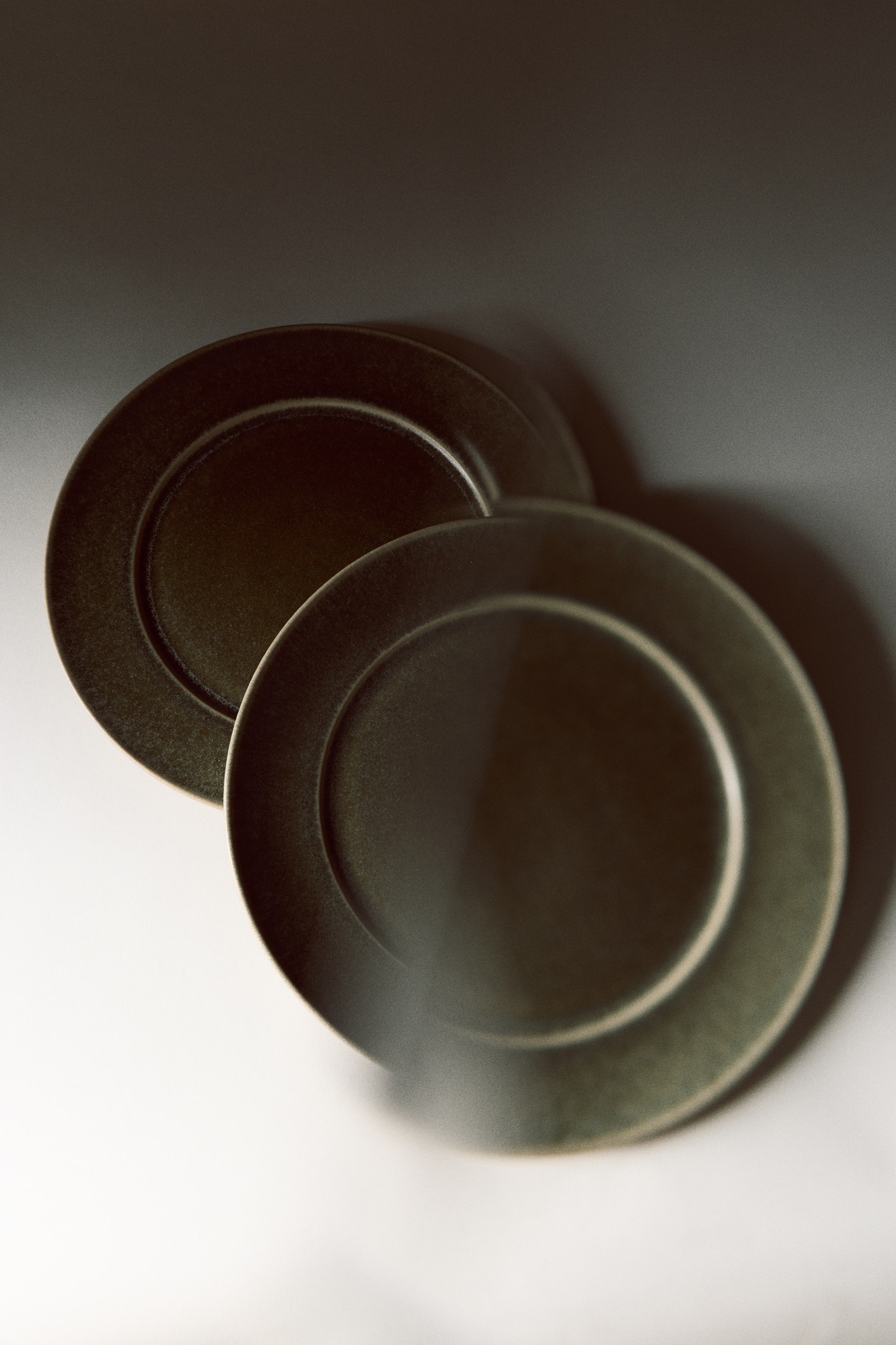 View larger image: Two dark green stoneware dinner plates are overlapping in an overhead view, showcasing a classic broad rim and a glossy reactive glaze finish with subtle speckling.