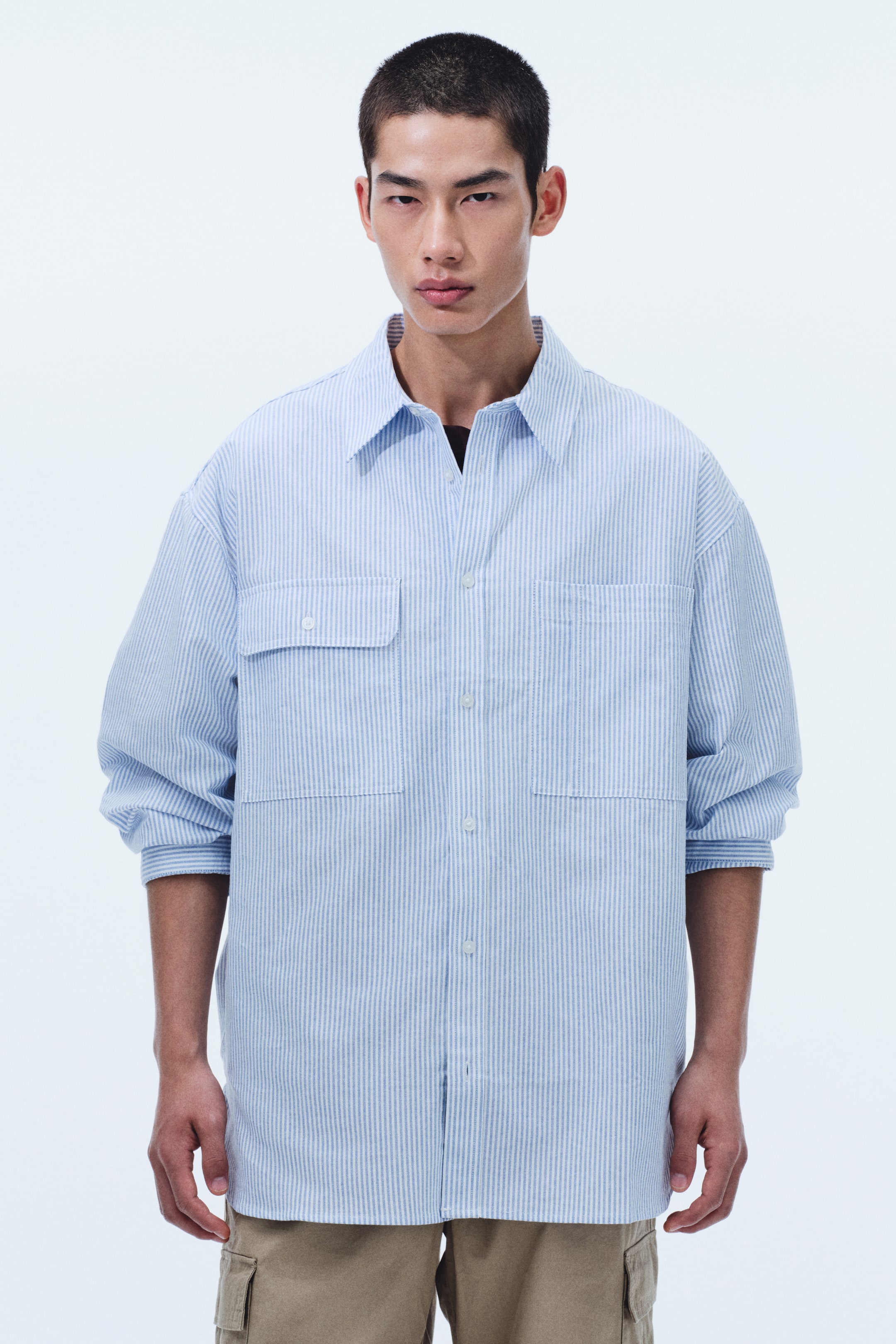 拡大画像を表示: ライトブルーとホワイトのストライプシャツは、スプレッドカラー、白いボタンの前開き、胸にボタン付きフラップの大きなパッチポケットが2つ付いているのが特徴です。長袖はロールアップされ、ダークカラーのTシャツの上にオープンで羽織るように着用され、オーバーサイズのシルエットに見えます。
