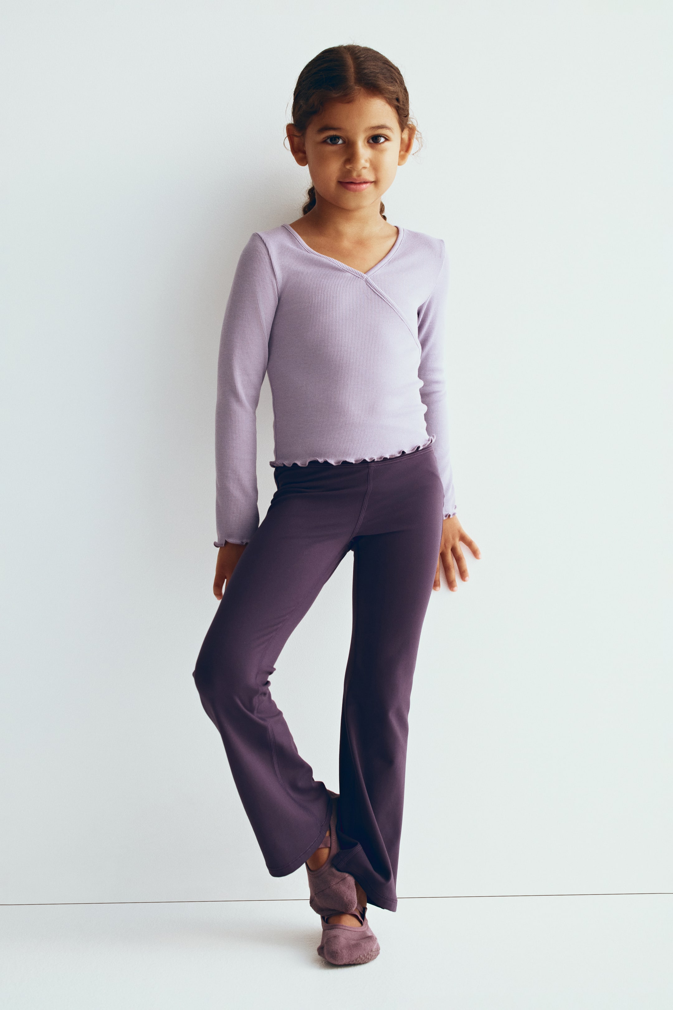 View larger image: A regular-fit activewear top in light purple ribbed knit is shown from the front, featuring a V-shaped neckline with a sewn-in wrap front. The long-sleeve top showcases overlocked lettuce-edge detailing on the cuffs and hem.