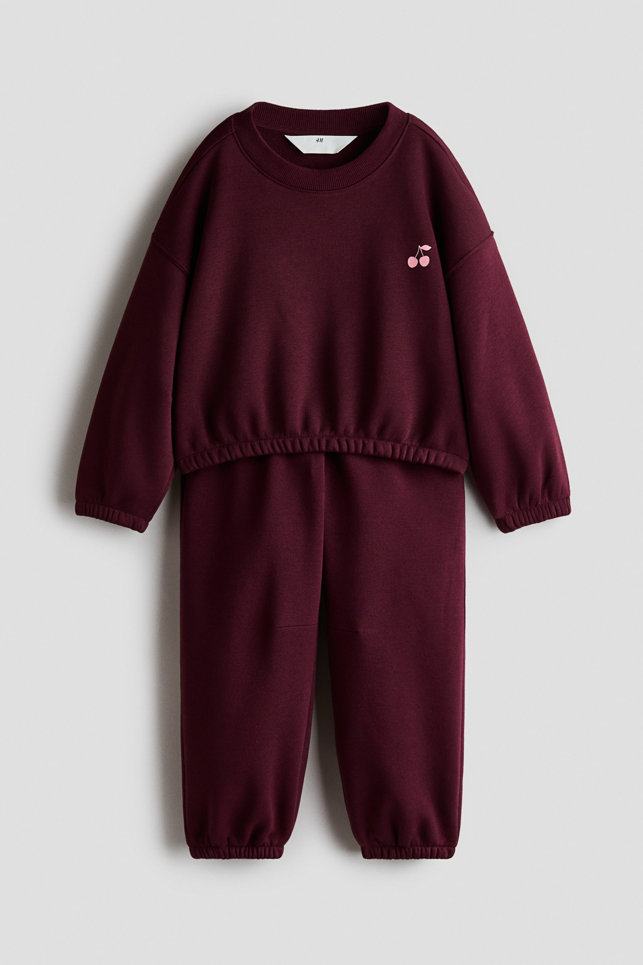 Se større billede: Et bordeaux todelt fleecesæt ses forfra og består af en langærmet sweatshirt og matchende joggingbukser. Sweatshirten har en ribstrikket rund hals, sænkede skuldre, elastiske manchetter og kant samt et lille pink kirsebærmotiv på brystet, mens de matchende bukser har rynkede, elastiske ankelmanchetter.
