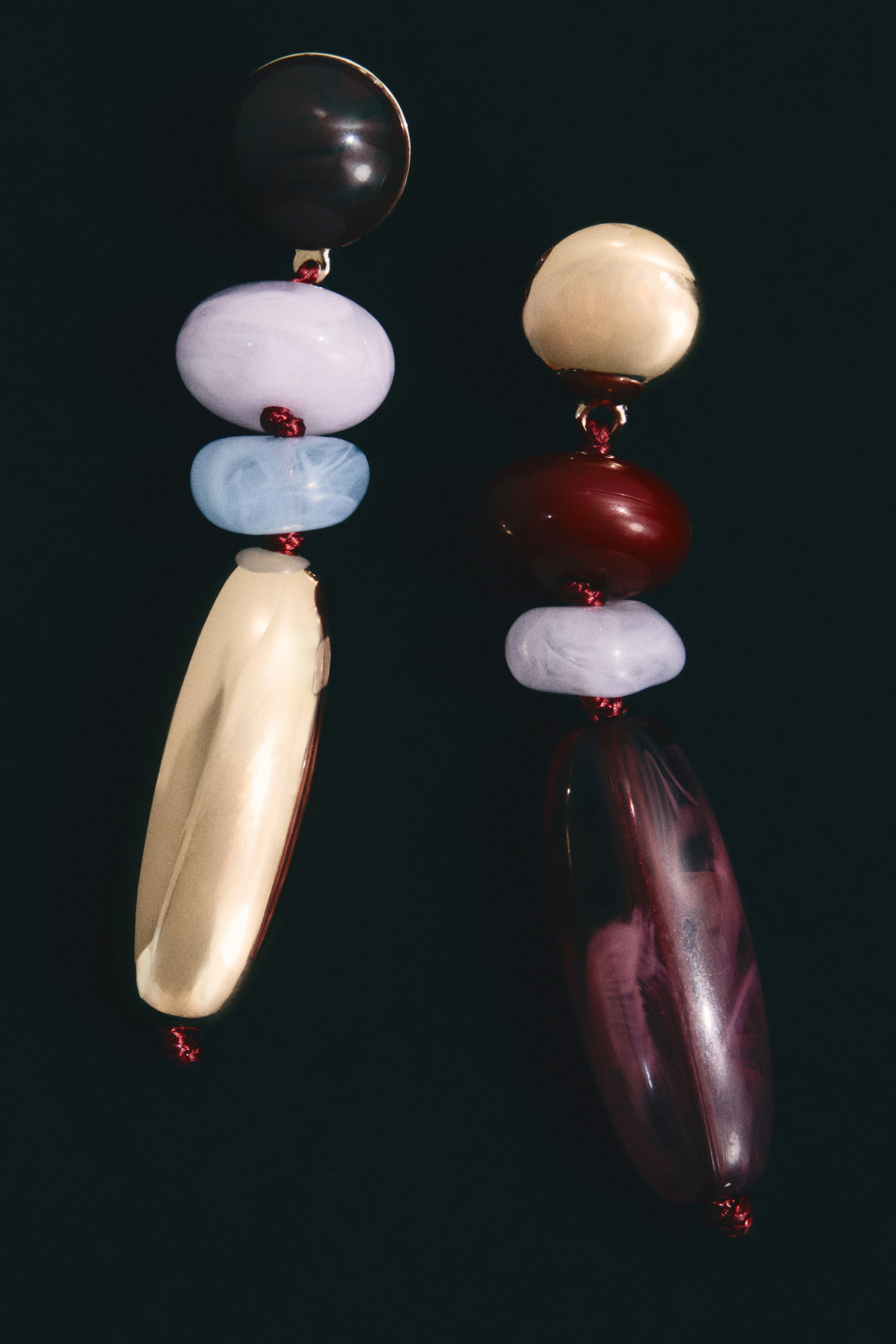 拡大画像を表示: 2つのマルチビーズ・ダングルイヤリングが垂直に垂れ下がっており、様々な不透明および半透明のビーズが赤いコードで接続されています。左のイヤリングには、ダークブラウンのドーム型ビーズ、続いてライトパープルとライトブルーの不規則な楕円形ビーズがあり、最後に光沢のあるゴールドの細長い楕円形ビーズで終わっています。一方、右のイヤリングには、ゴールドのドーム型ビーズ、ダークレッドの不規則な楕円形ビーズ、ライトパープルの不規則な楕円形ビーズ、そして半透明のダークパープルの細長い楕円形ビーズが見られます。