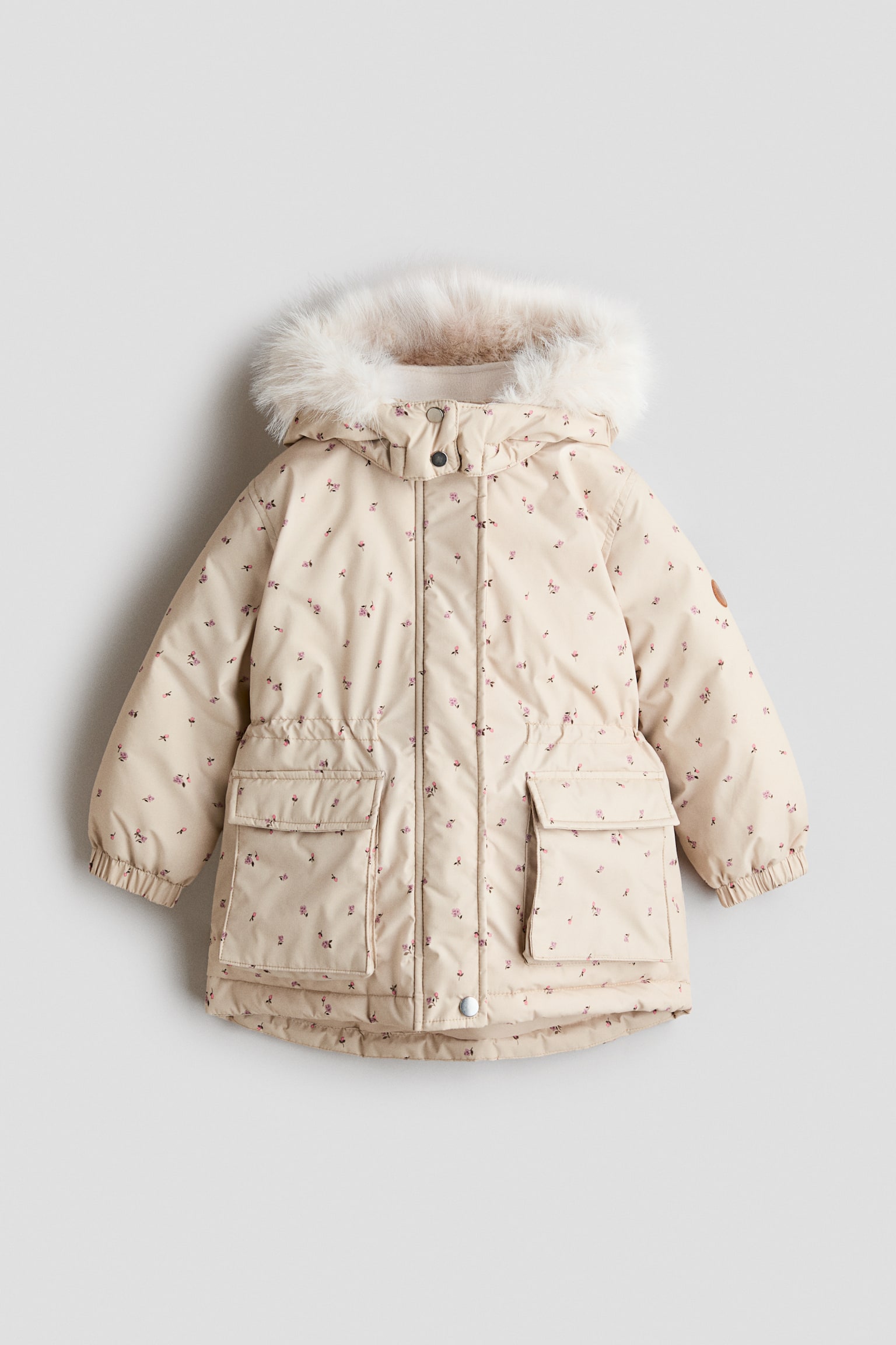 En vadderad vinterjacka för barn visas framifrån och framhäver ett ljusbeige tyg med ett fint, över hela plagget spritt rosa blommönster. Jackan har en huva kantad med fluffig krämfärgad fuskpäls, en dold dragkedja framtill med tryckknappar, en resår i midjan och två stora påsydda fickor framtill.