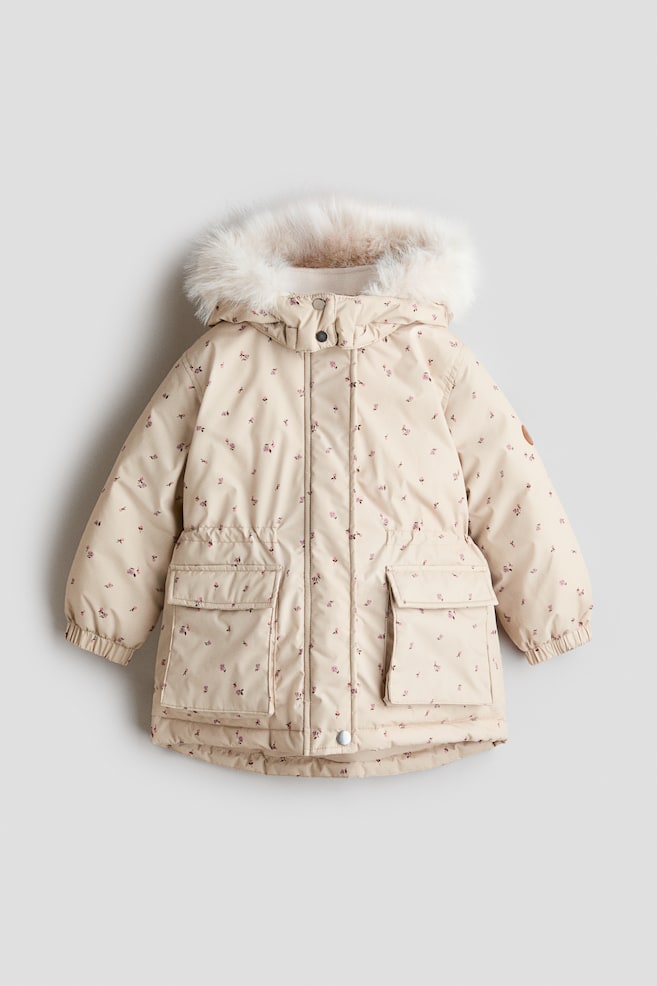 Un abrigo acolchado de invierno para niña aparece de frente, mostrando un tejido beige claro con un delicado estampado floral rosa integral. El abrigo presenta una capucha ribeteada con suave pelo sintético crema, un cierre frontal de cremallera oculta con botones a presión, cintura elástica y dos amplios bolsillos delanteros con solapa. - 1