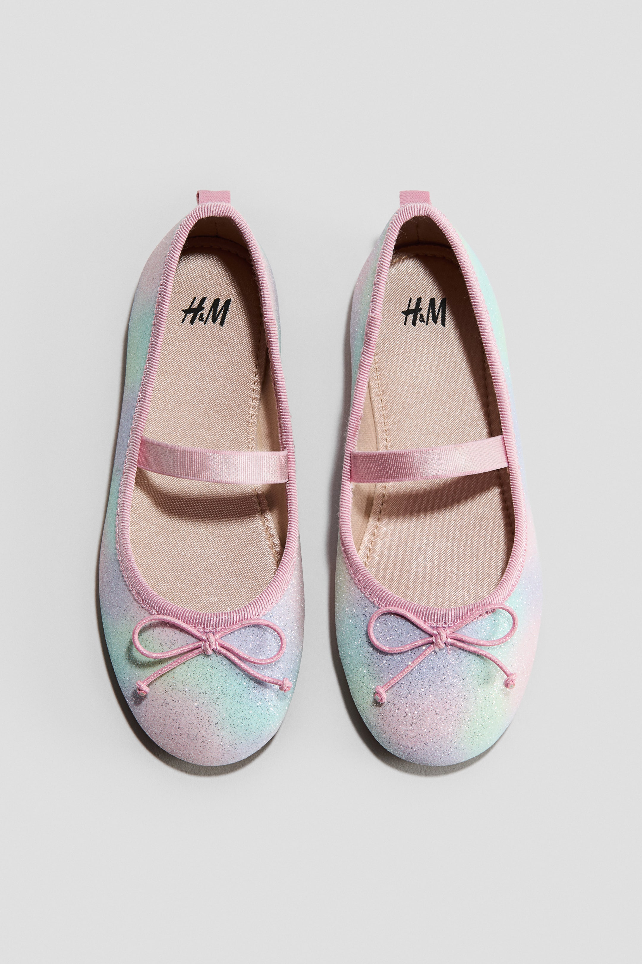 Voir une image plus grande: Une paire de ballerines scintillantes pour enfants présente un design ombré pastel, avec une bride élastique rose sur le cou-de-pied et un nœud en gros-grain rose à l'avant. Chaque chaussure est également dotée d'un liseré rose côtelé autour de l'ouverture, d'une petite languette rose à l'arrière et d'un logo H&M sur la semelle intérieure.