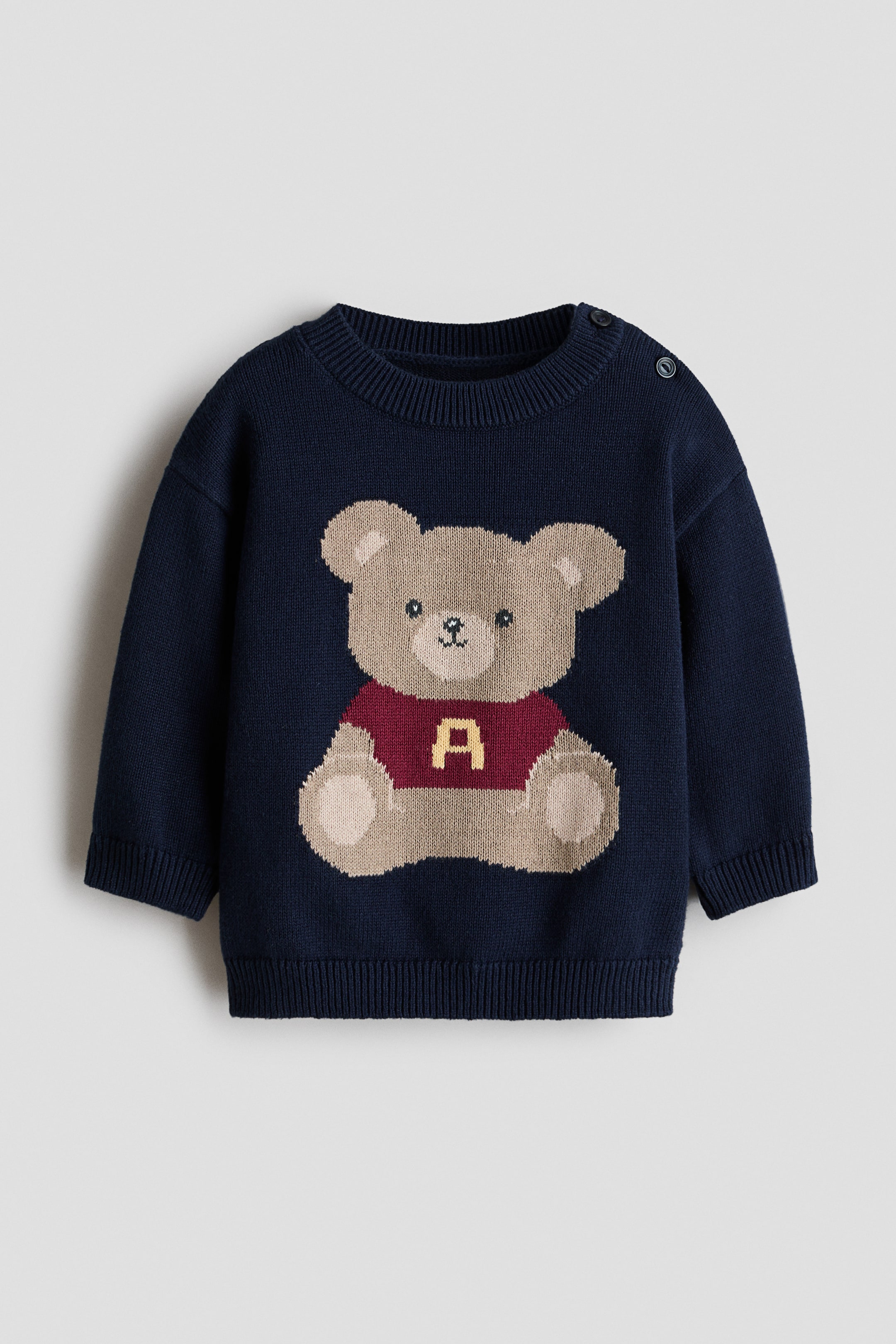 Grotere afbeelding bekijken: Een zachte, jacquard gebreide katoenen trui met een groot, lichtbruin teddybeermotief op de voorkant, gekleed in een bordeauxrode top met een gele 'A'. Het ontwerp heeft een ronde halslijn met ribboord, twee knopen op de linkerschouder, verlaagde schouders, geribde manchetten en een geribde zoom.