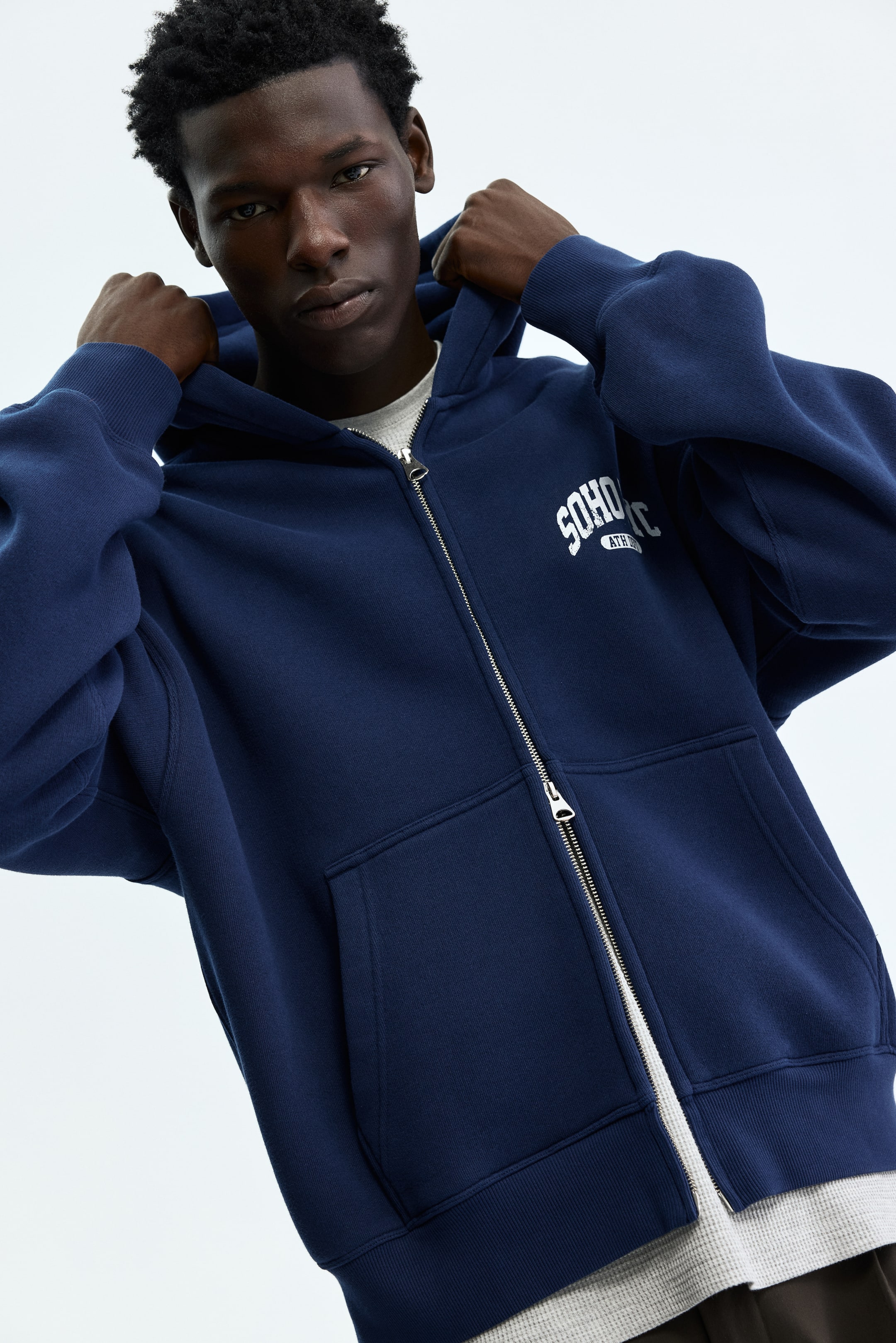 View larger image: A navy blue zip-up hoodie featuring a silver-toned zipper down the front, white 'SOHO ATHL' text on the left chest, and two front pouch pockets. The hoodie showcases long sleeves with ribbed cuffs and a ribbed hem, worn over a light gray thermal shirt.