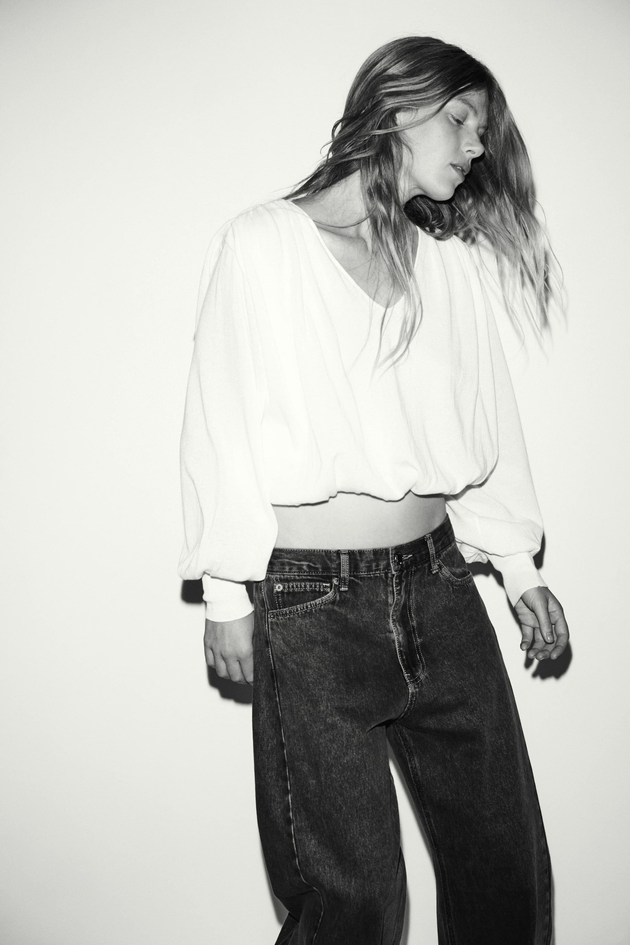 View larger image: A short, relaxed-fit white knit top featuring a V-neckline, long balloon sleeves with wide cuffs, and a bubble hem is worn by the model. The top is paired with dark-wash, wide-leg denim jeans.