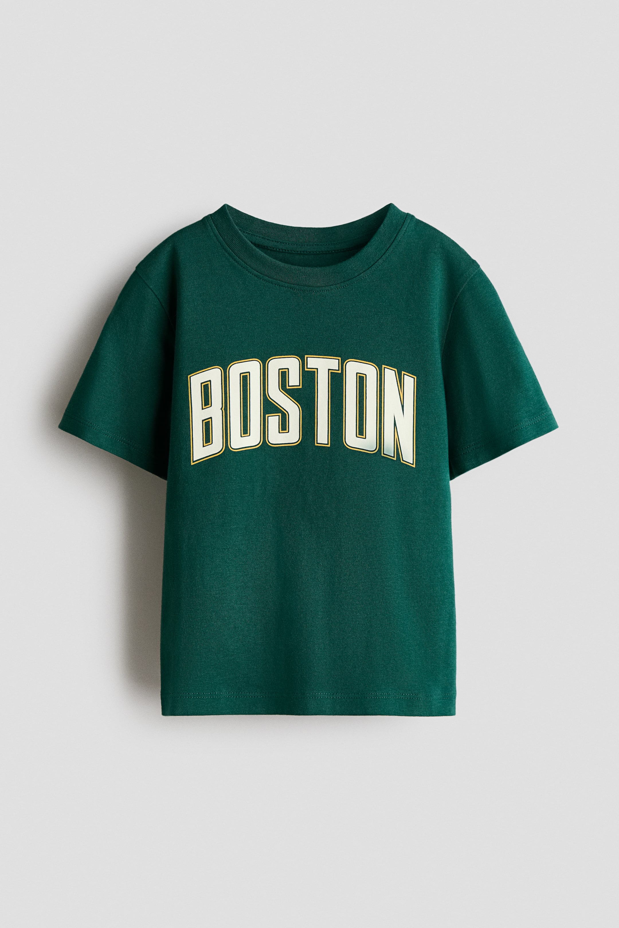 Xem ảnh lớn hơn: Một chiếc áo phông màu xanh lá đậm bằng vải jersey in cotton đang trình bày mặt trước của nó, có đặc điểm cổ tròn và tay ngắn. Mặt trước đang trưng bày từ "BOSTON", hiện lên bằng chữ in hoa lớn, cong, màu kem với đường viền màu vàng trên ngực áo.