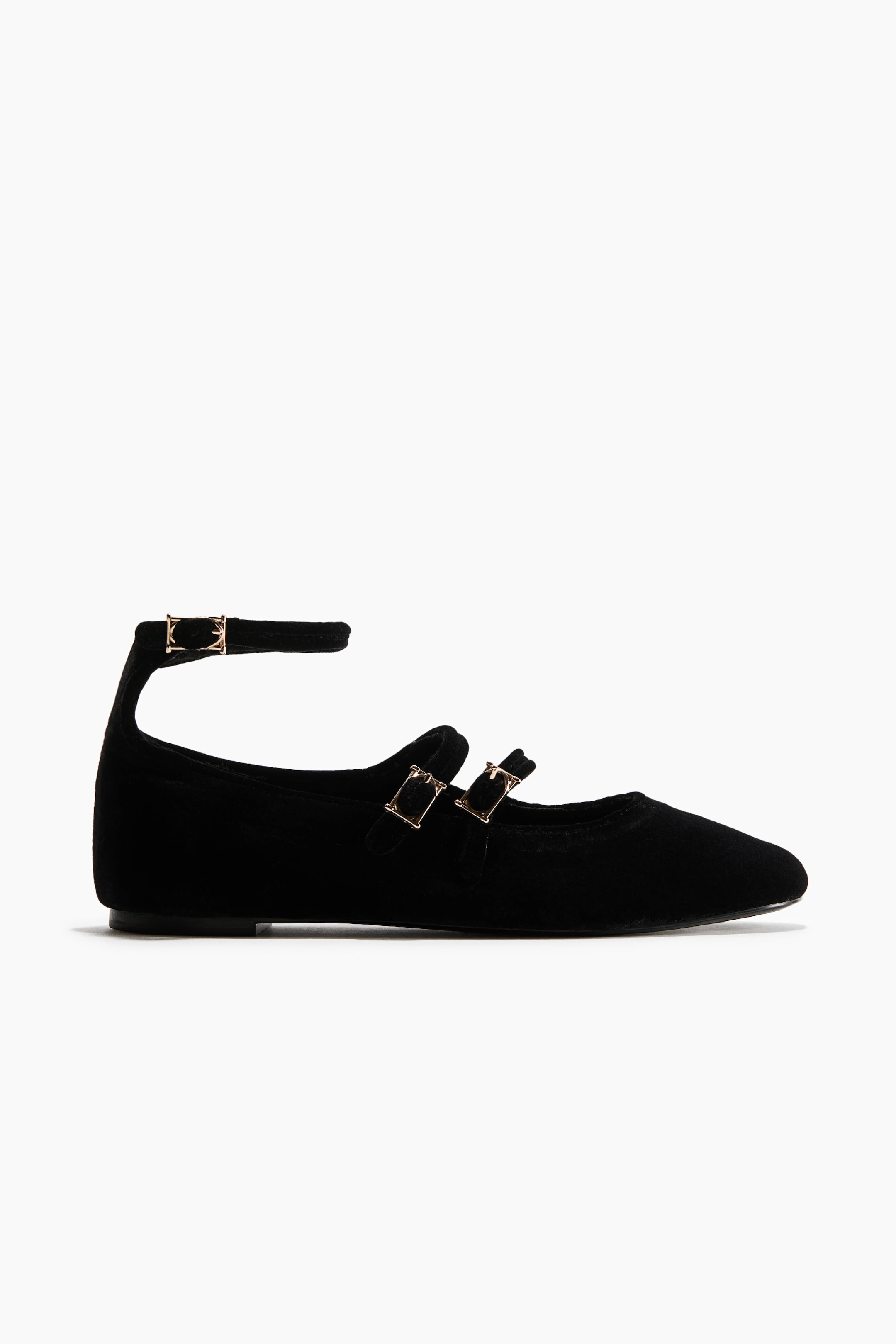 View larger image: A black velvet ballet flat is shown from a side profile view, featuring a rounded toe and a low, flat sole. The design includes two narrow, adjustable straps running across the top of the foot and a third strap wrapping around the ankle, all secured with small, gold-tone metal buckles.