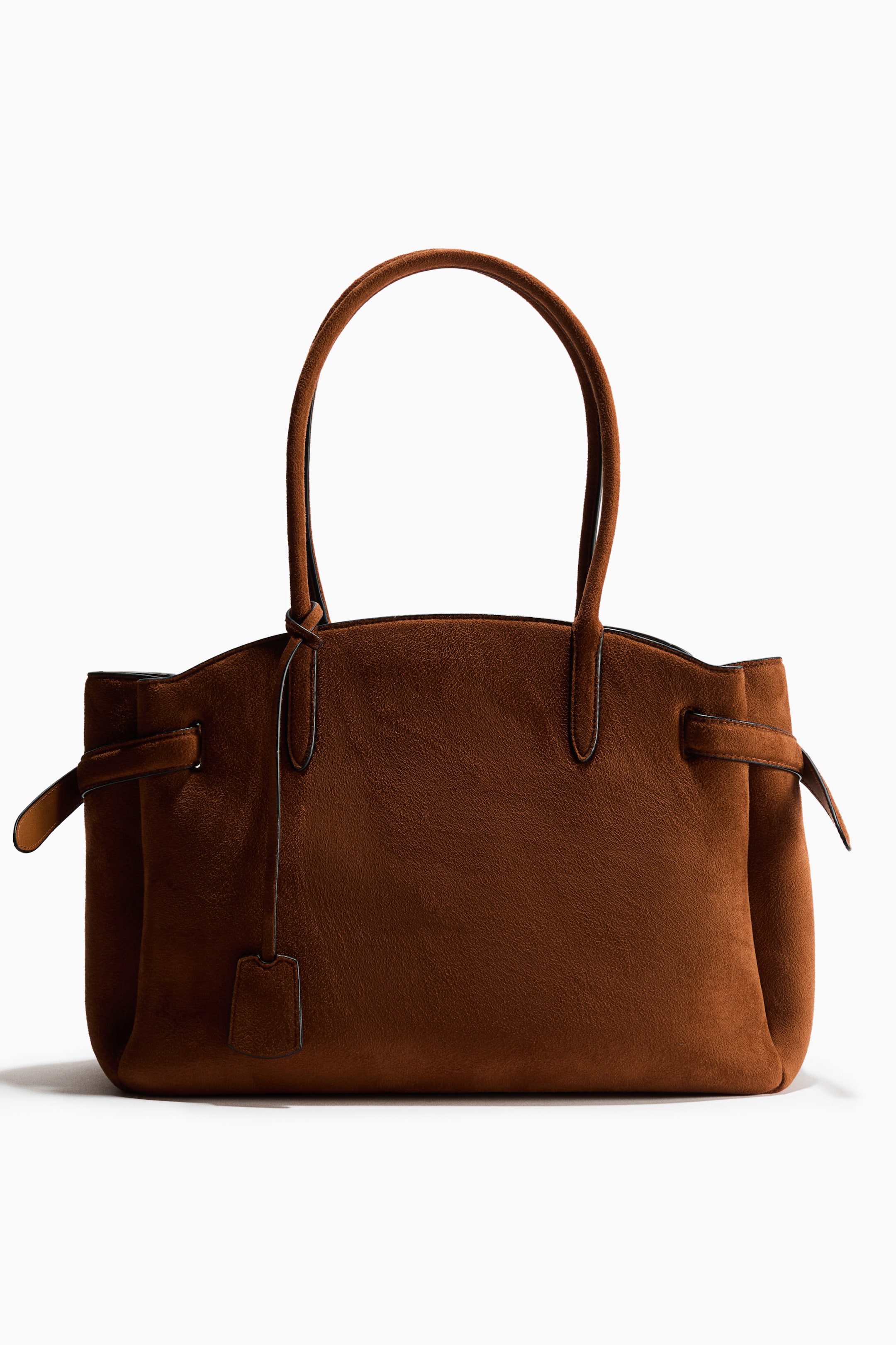 View larger image: Front-facing brown textured tote bag with long top handles. A decorative, detachable bag tag hangs from one handle, and crossover straps are fastened with a stud on each short side, pulling the top edges inward.