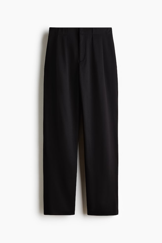 Black tailored trousers are shown from the front, featuring a clean waistband with front pleats and a concealed fly closure. The trousers are designed with slightly tapered legs that have a subtle crease running down the centre. - 1