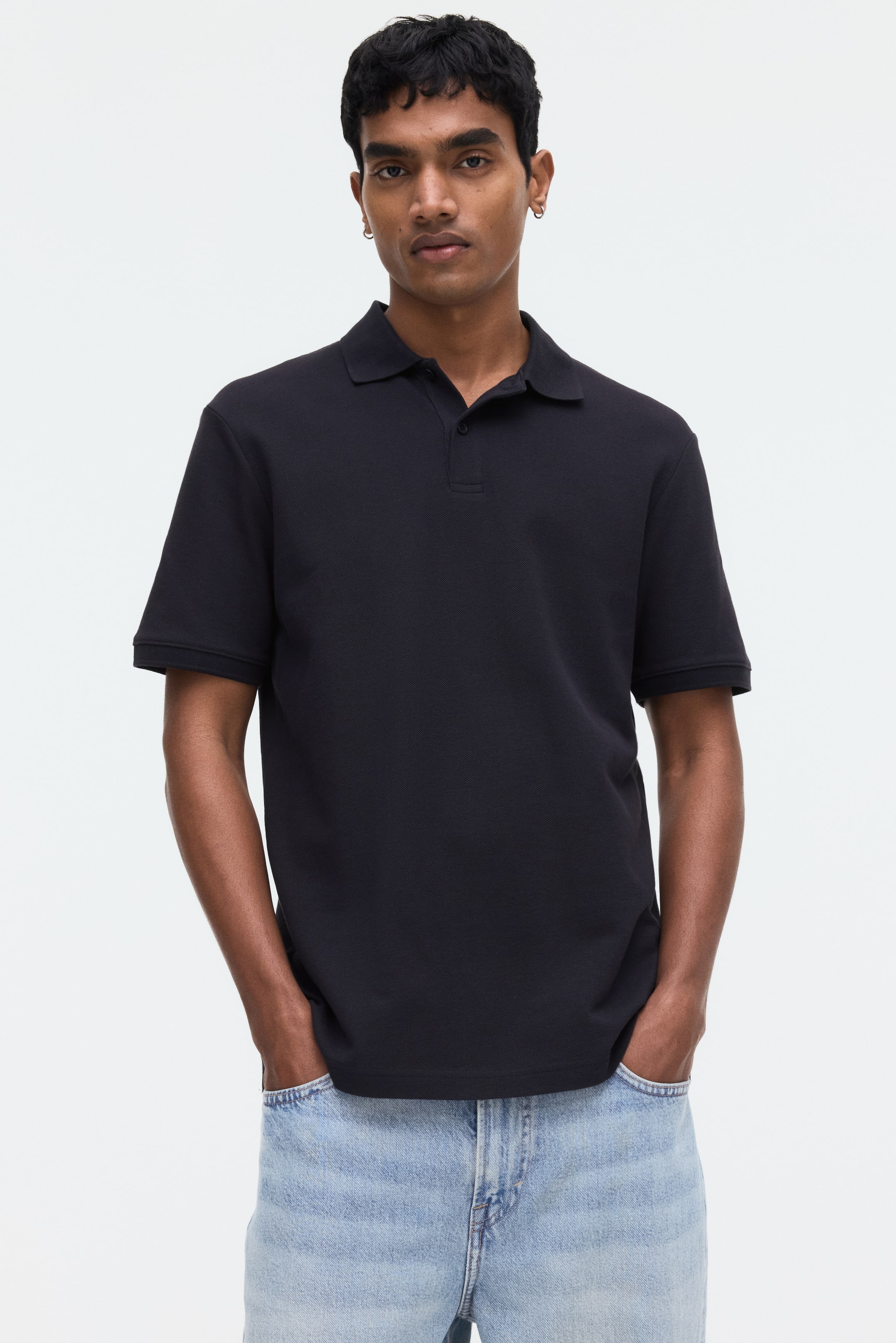 Größeres Bild ansehen: Ein schwarzes Regular Fit Polo-Shirt mit kurzen Ärmeln, einem Kragen und einer Zwei-Knopf-Leiste wird vom Model getragen, das auch eine hell gewaschene Jeans trägt.