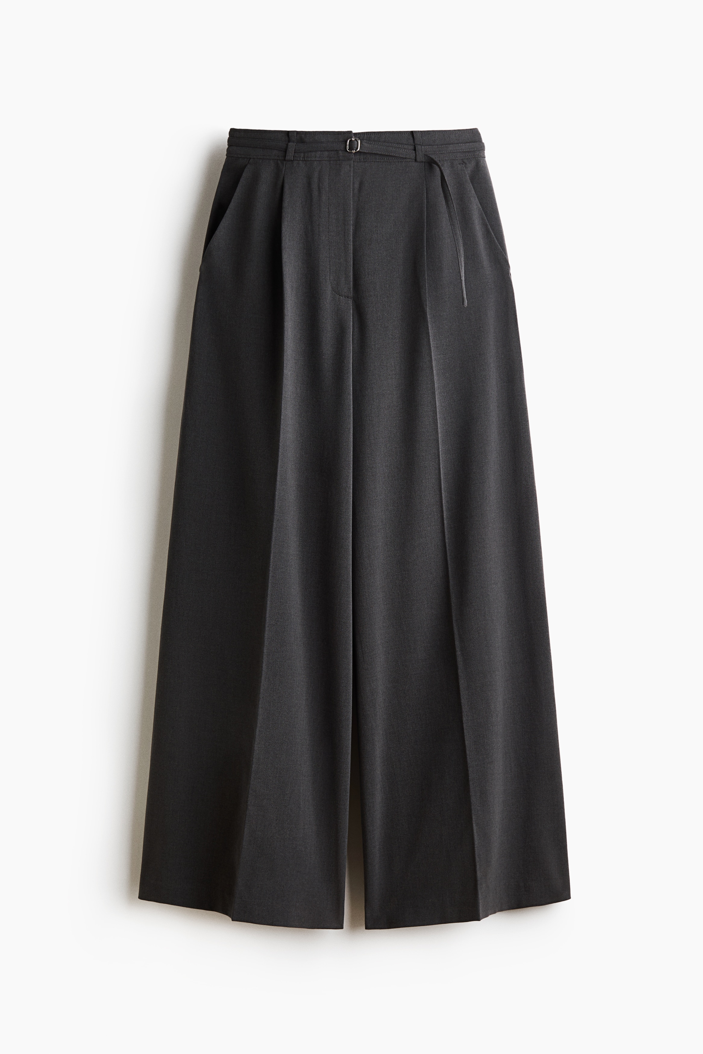 Pantaloni croiți cu croială largă și talie înaltă, din material țesut gri închis, sunt purtați de model, care stă cu o mână sprijinită într-un buzunar lateral oblic. Pantalonii au dungi călcate pe față și o curea îngustă asortată, fiind stilizați cu un top alb cu mânecă lungă, băgat în pantaloni.
