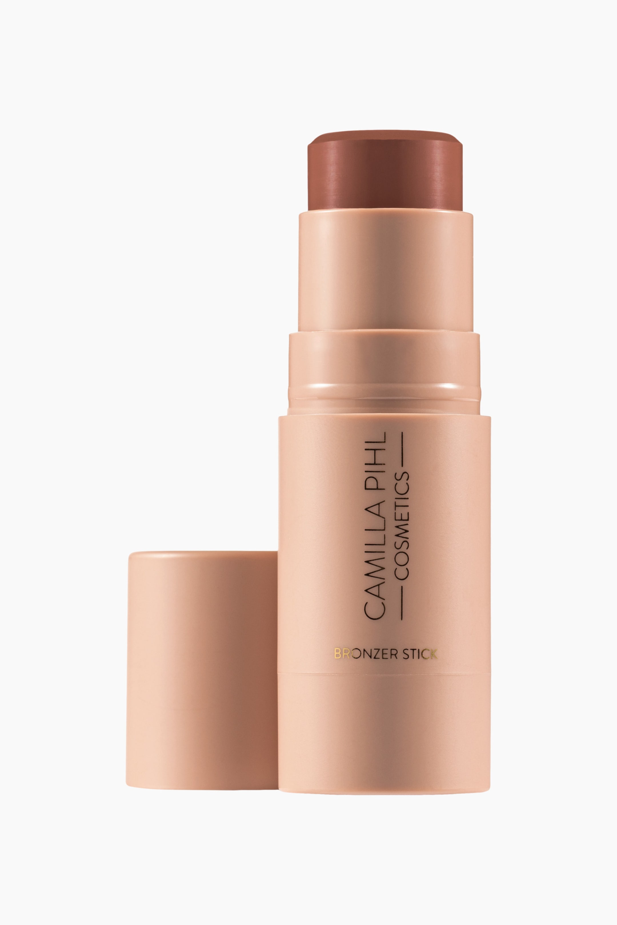 Visa större bild: En bronzerstick står upprätt, något riktad åt höger, med sitt ljuspersikofärgade matta hölje som visar "Camilla Pihl Cosmetics" och "Bronzer Stick" i guld. En medelbrun rundad bronzerspets sticker ut från toppen, och dess matchande ljuspersikofärgade lock vilar till vänster om sticket.