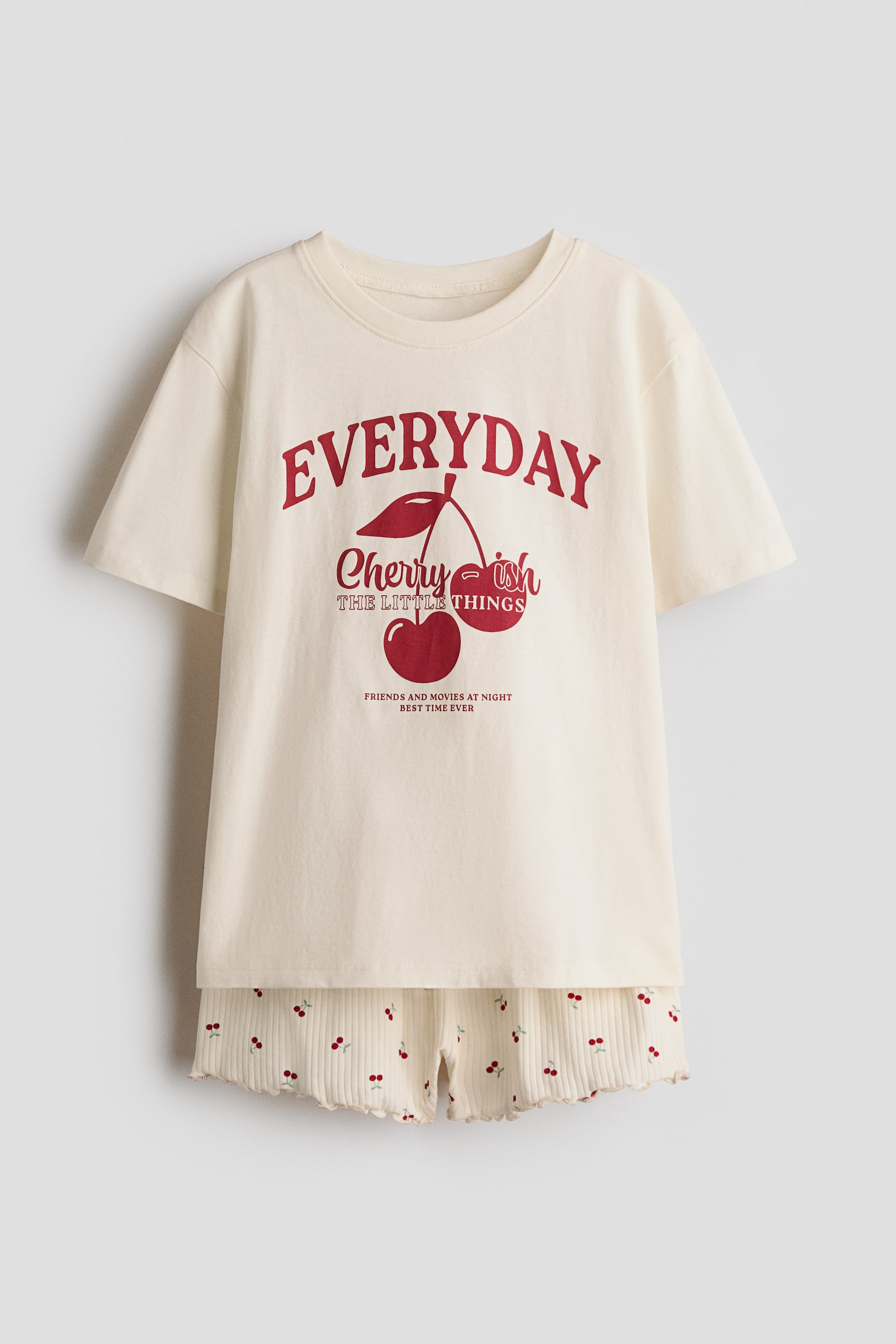 Visualizza l'immagine ingrandita: Un set pigiama è mostrato frontalmente, composto da una t-shirt color crema con scollo rotondo, orlo dritto e una stampa grafica rossa "EVERYDAY Cherry ish THE LITTLE THINGS" con due ciliegie. Sotto sono stratificati shorts a costine color crema, con vita elasticizzata, orli arricciati e un motivo a ciliegie rosse su tutta la superficie.