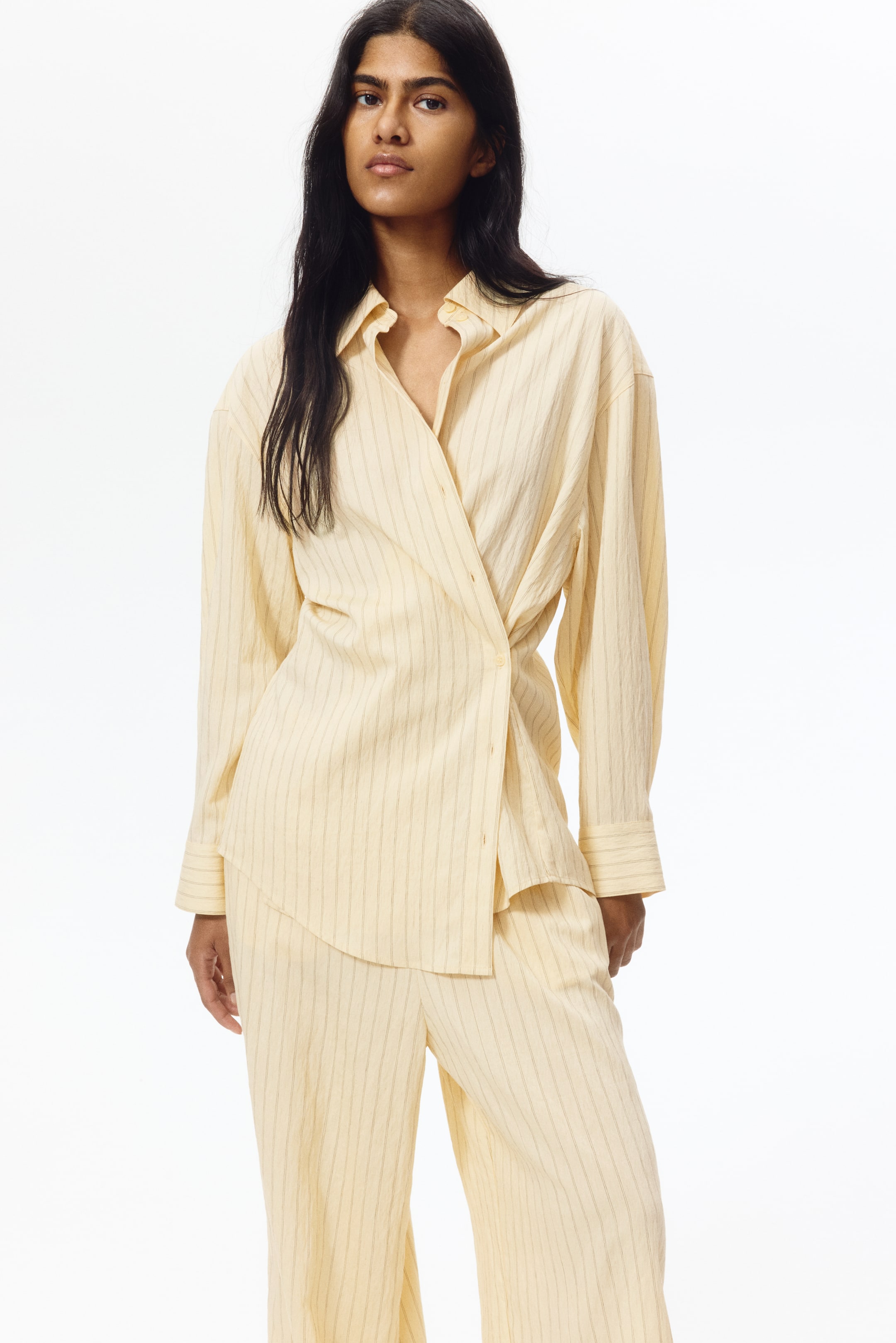 View larger image: A cream-coloured, pinstriped shirt featuring a spread collar, long sleeves with buttoned cuffs, and an asymmetric wrap-style front closure. Matching wide-leg trousers complete the set, with both garments displaying thin vertical pinstripes.