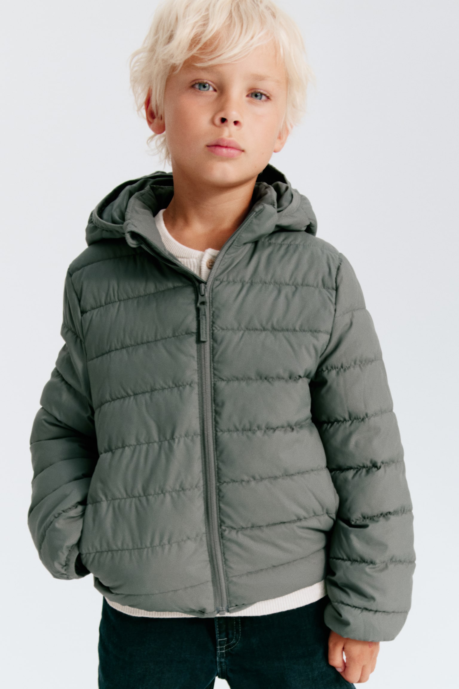 A muted green quilted puffer jacket is facing forward, featuring a full-length zip closure, a stand-up collar, and a hood with elasticated trim. It is displaying elasticated cuffs and hem. - 1