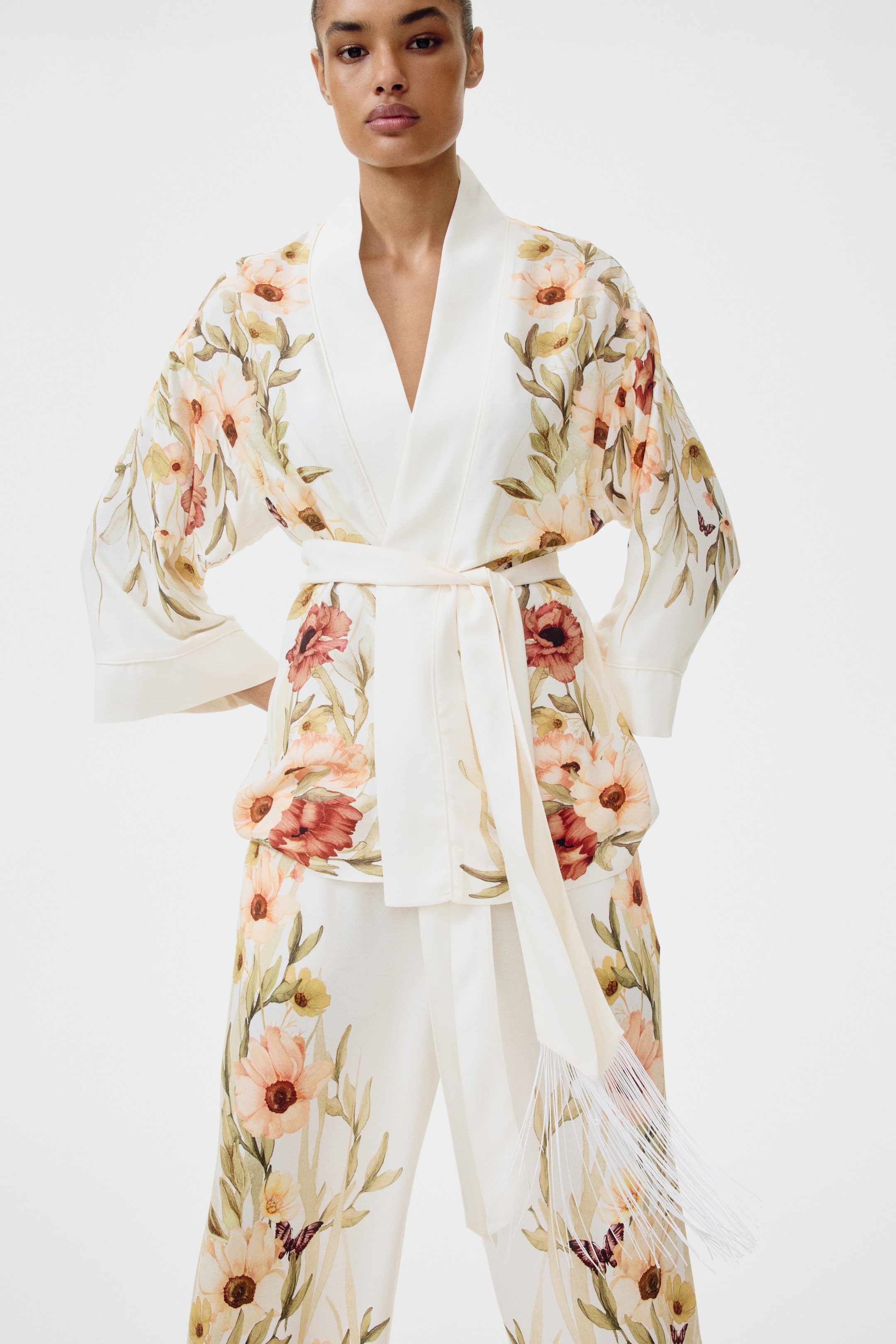 Voir une image plus grande: Un ensemble deux pièces crème, présenté de face, comprend une veste style kimono et un pantalon droit assorti, tous deux ornés d'un imprimé floral aquarelle dans des tons de pêche, rouge et vert. La veste est conçue avec un large col châle bordé de satin et se noue à la taille avec une ceinture à nouer assortie, dont une extrémité est agrémentée de franges blanches.